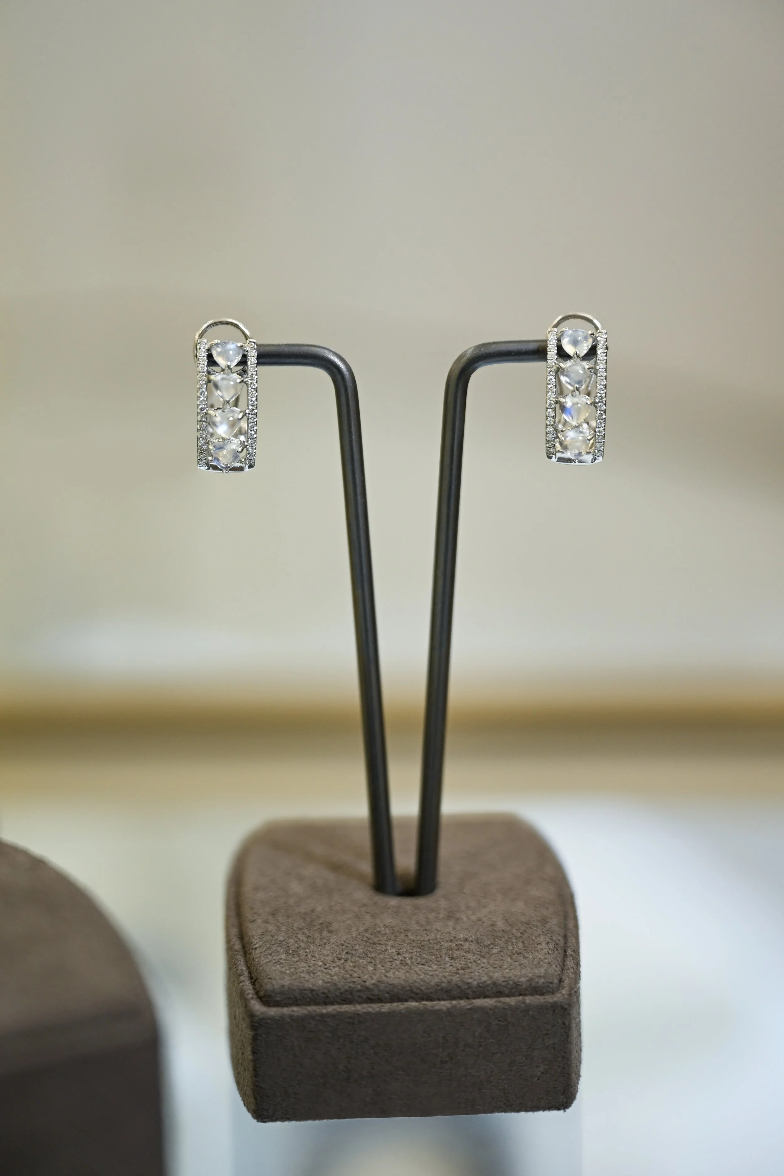 A pair of silver earrings with clear gemstones hanging from hooks, displayed on a black stand on a beige surface.