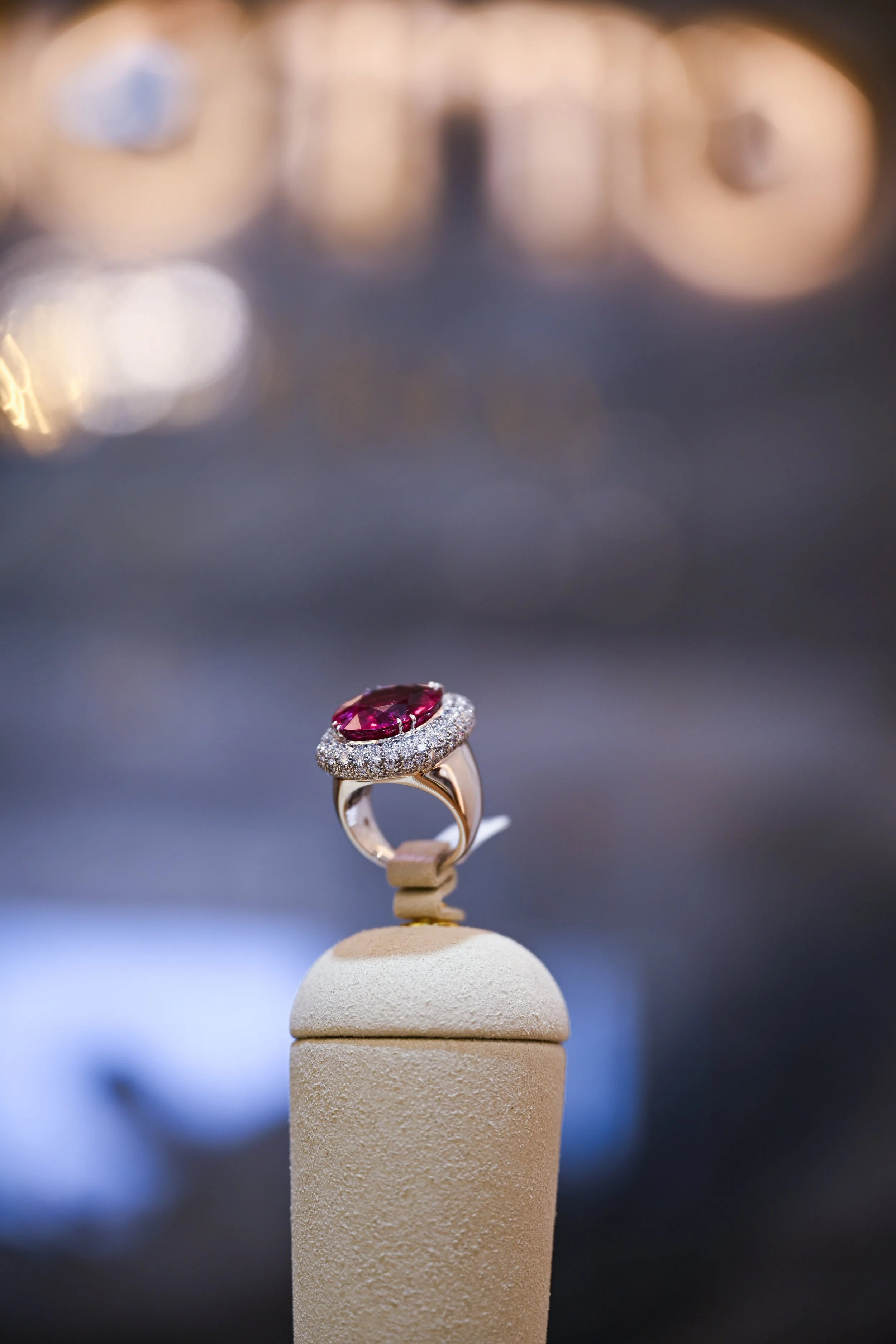 Close-up of a fancy ring with a large pink gemstone surrounded by small diamonds, displayed on a beige ring holder, with a blurred background of golden lights.