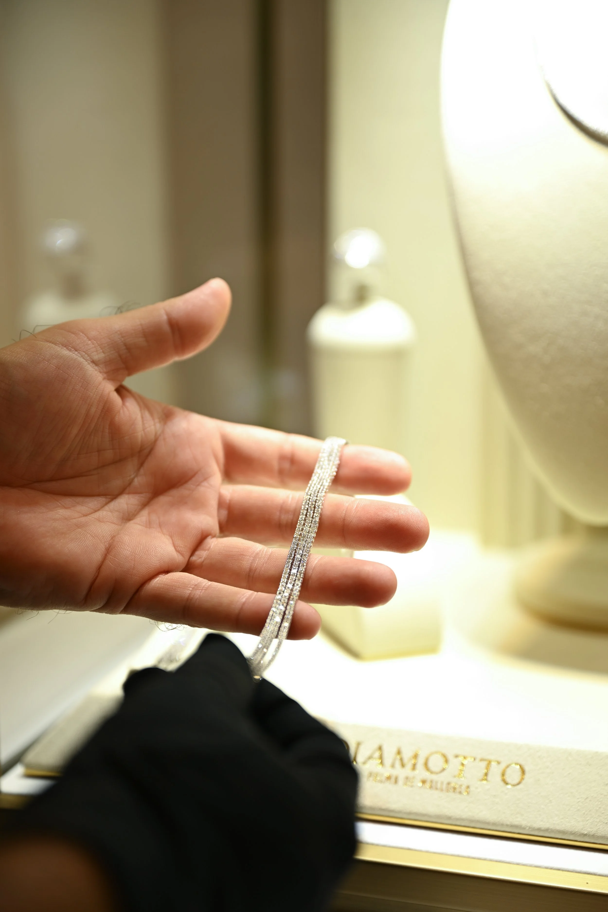 A person's hand holding a thin, sparkling silver bracelet in front of a display case with a jewelry box inside. The jewelry box is gold and silk-lined, with some text visible on it.
