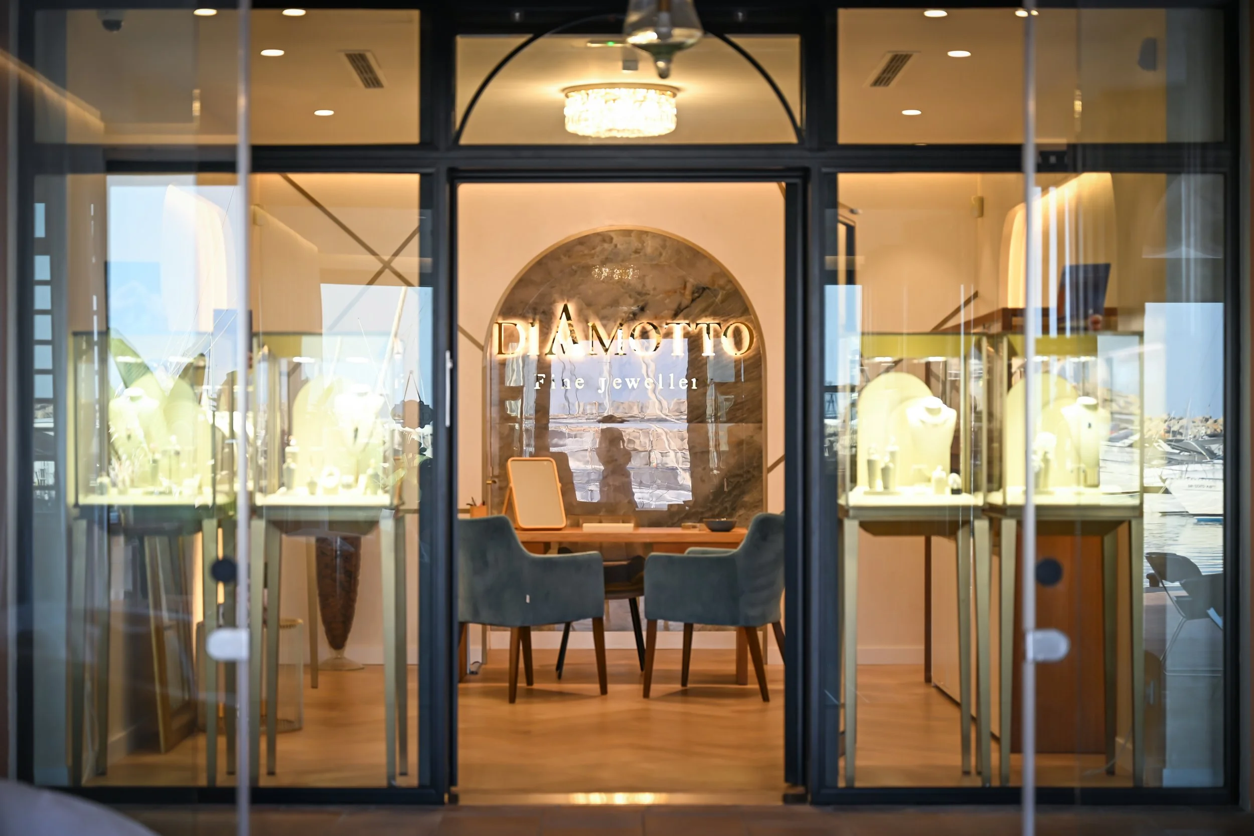 View of jewelry store window with illuminated sign that reads 'DIAMTTO Fine Jeweller',  in Puerto Portals, showcasing jewelry displays inside, chandelier hanging from ceiling, and reflection of street outside.