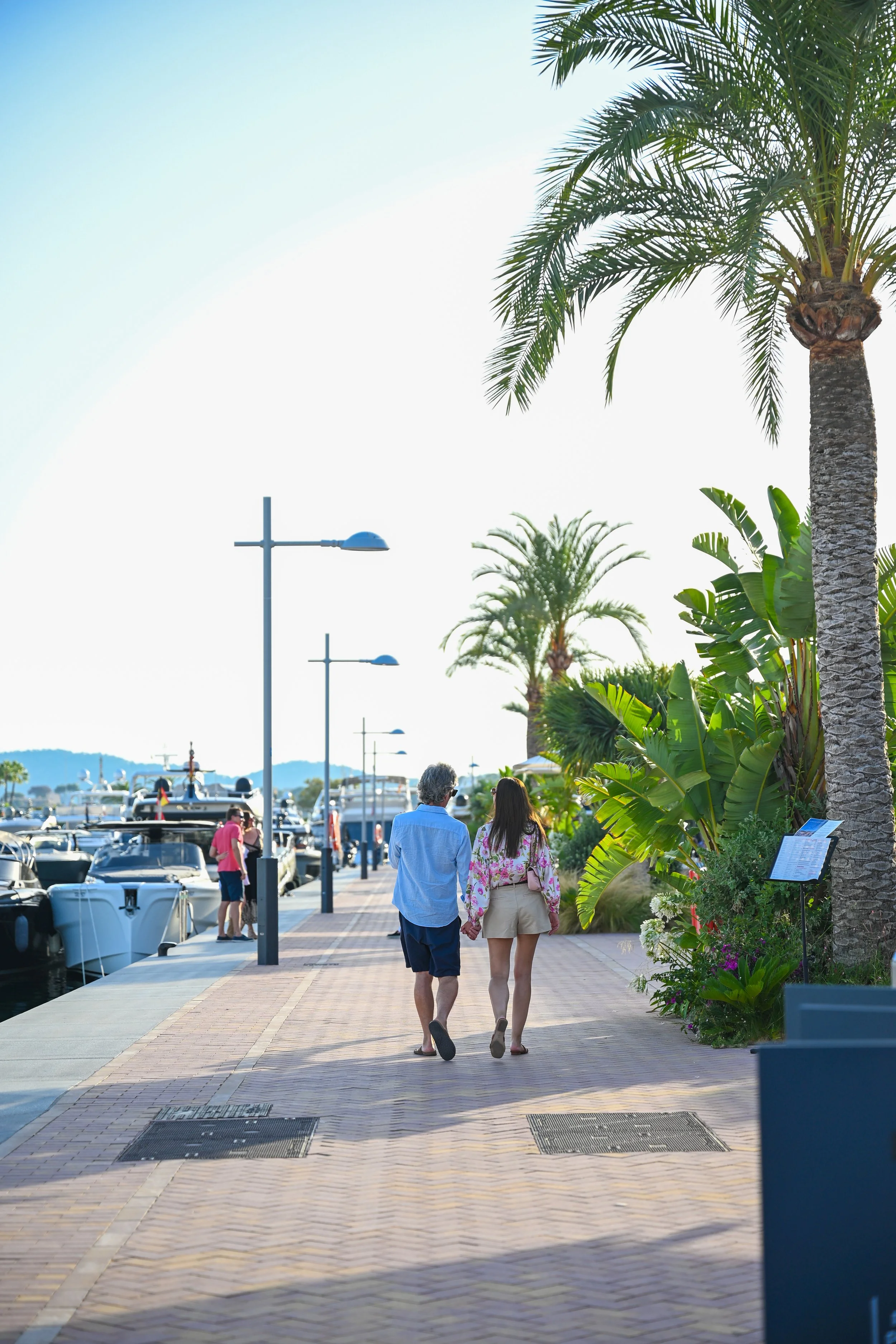 A couple walking hand in hand along a waterfront promenade lined with palm trees and street lamps, with boats docked in the marina and a blue sky overhead.