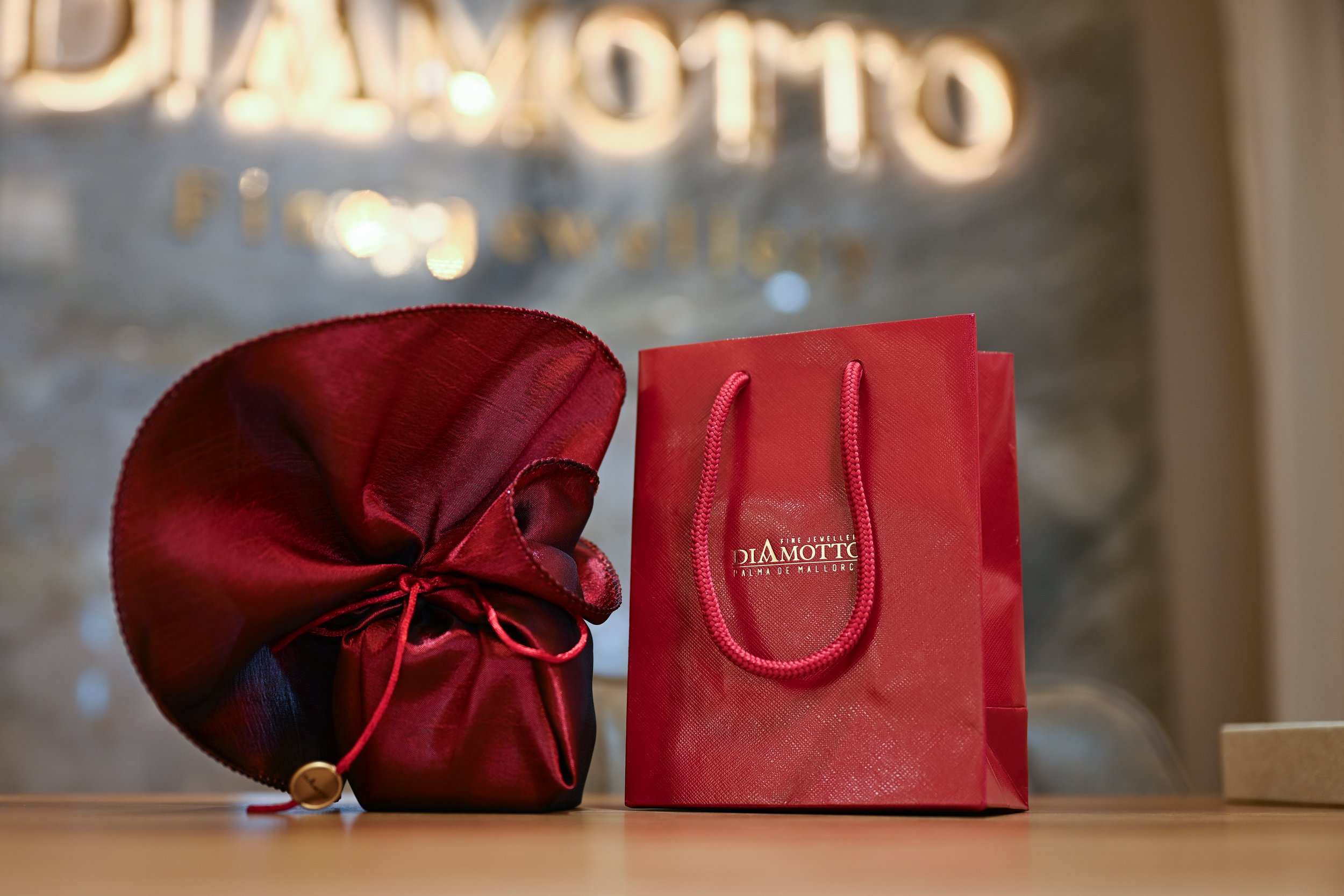 A red fabric gift bag and a red satin drawstring pouch placed on a wooden surface with a background featuring blurred shiny gold letters.