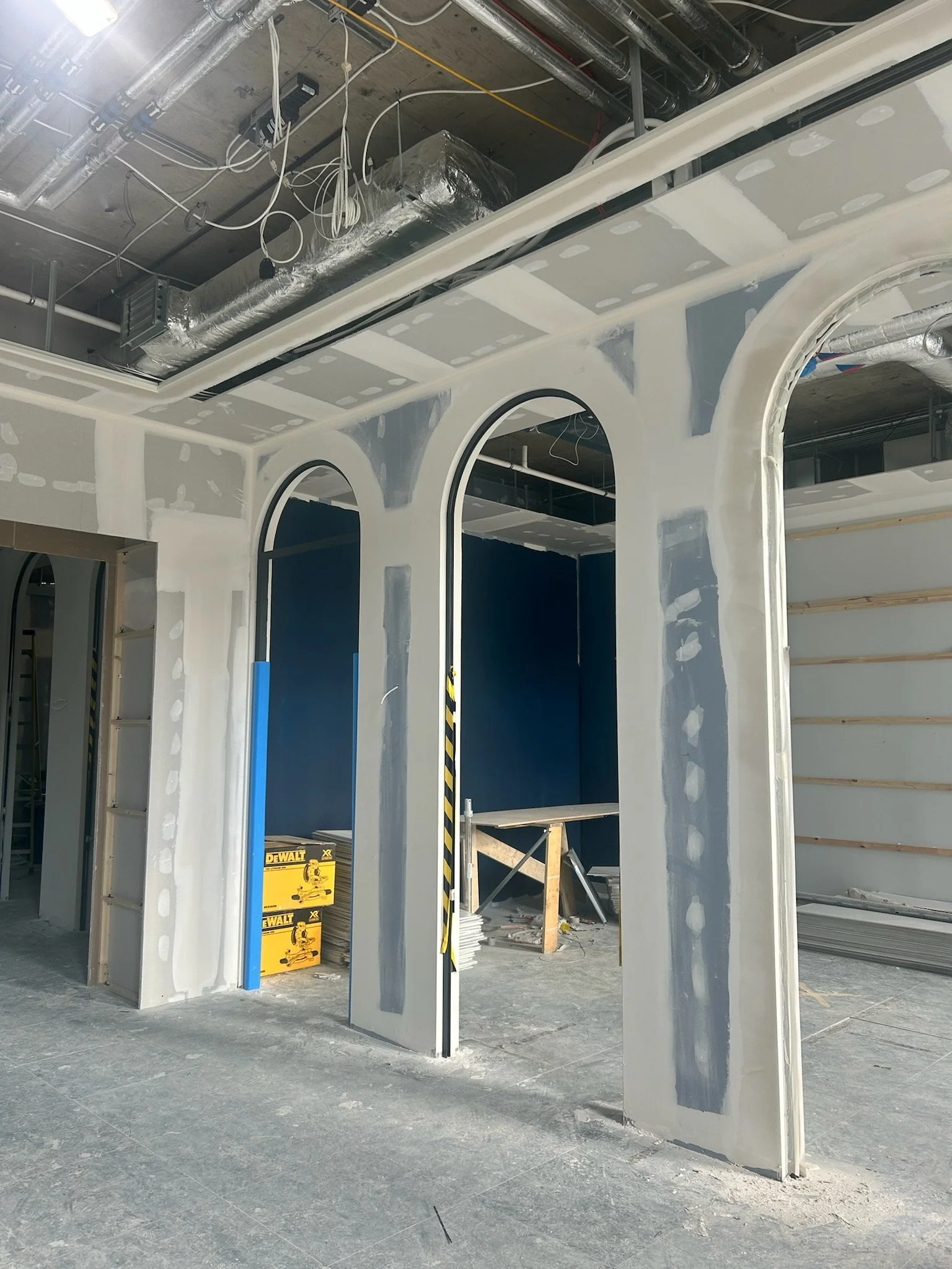 Interior of a building under construction, with drywall partially installed, electrical wiring hanging from the ceiling, and construction tools such as a DEWALT boxed power tool on the floor.