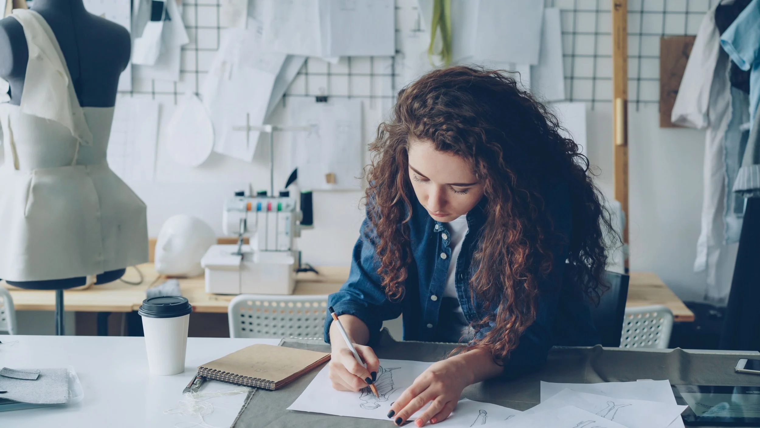 Young woman with curly hair sketching fashion designs at a worktable in a fashion design studio with mannequins and fabric patterns.