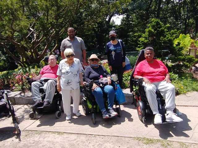 A group of people, some in wheelchairs, outdoors in a garden setting with trees and flowers in the background.