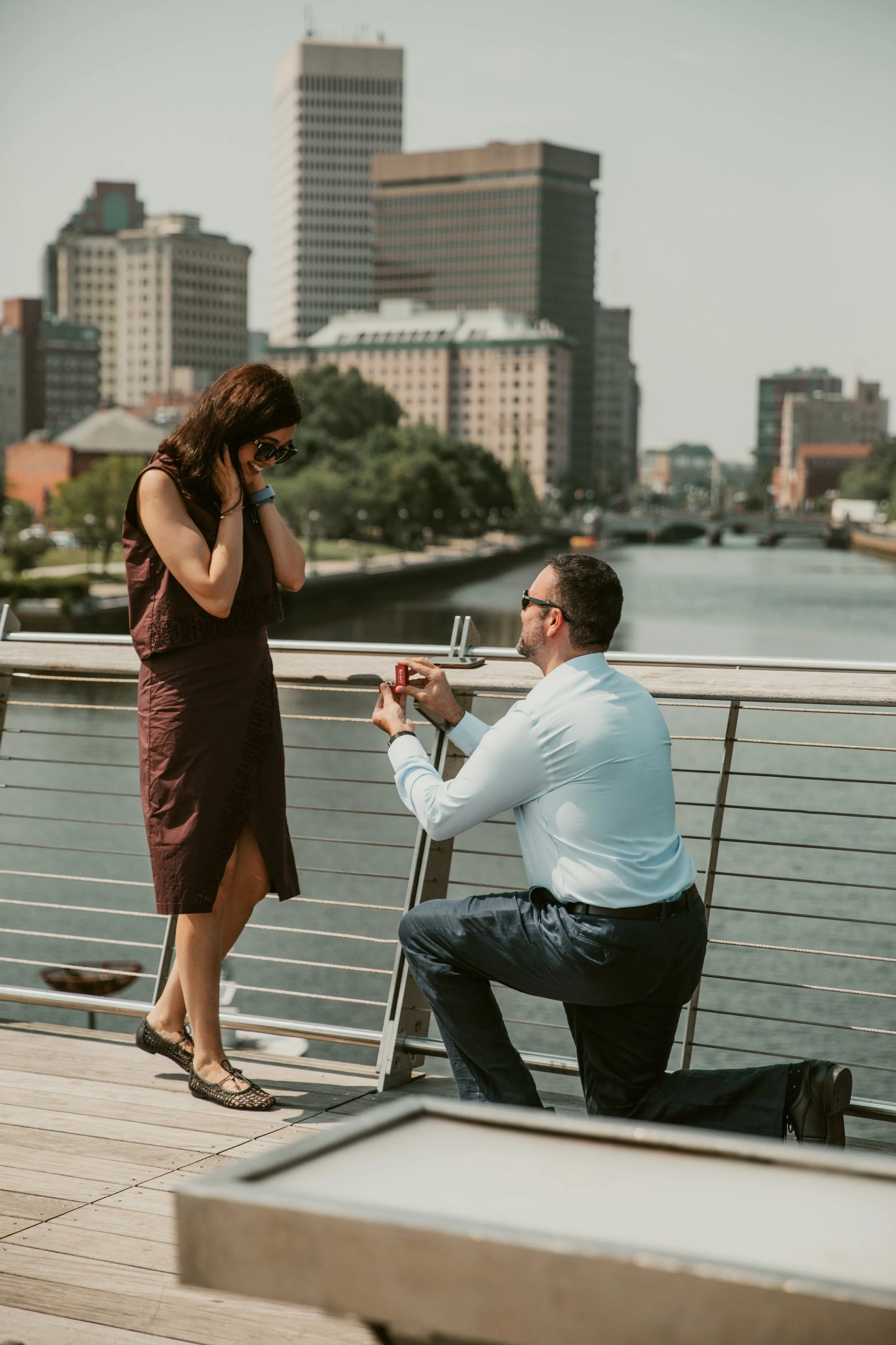 A man is kneeling on one knee, proposing with a ring to a woman on a bridge with a city river and skyline in the background.