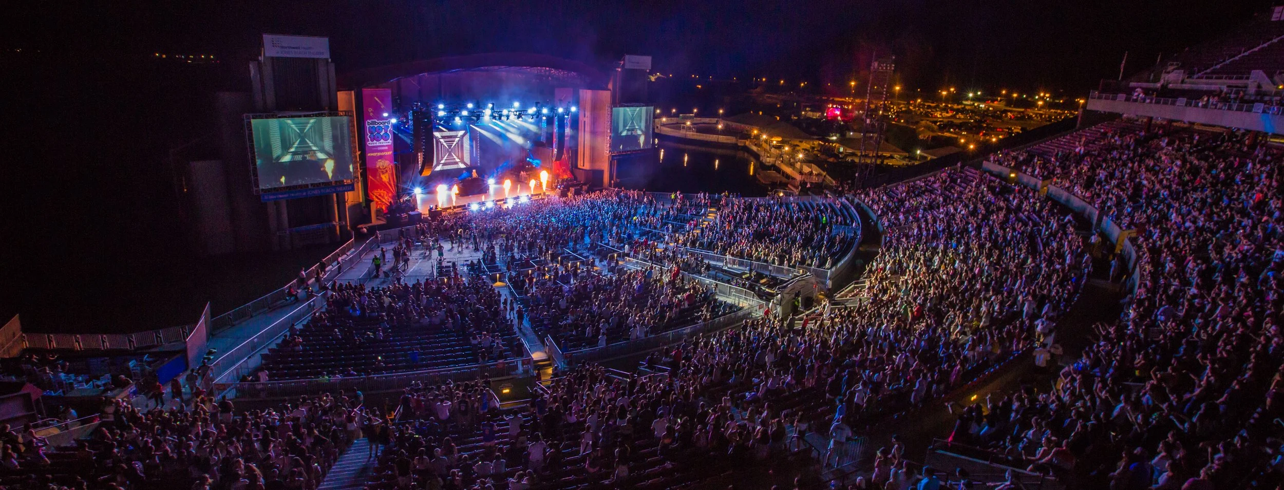 Nighttime outdoor concert at a large stadium with a stage, bright lights, and a crowd of people.
