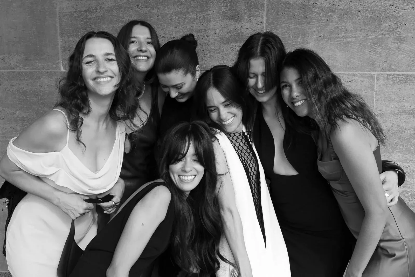 A group of seven women smiling and hugging each other against a textured wall."}