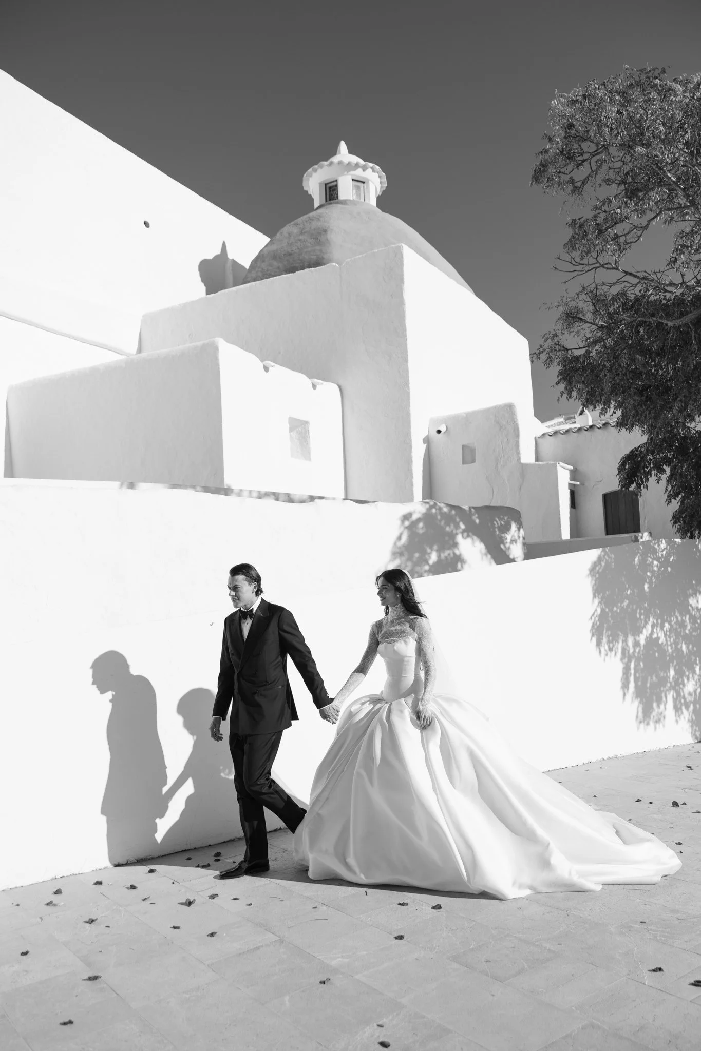 Black and white photo of a bride and groom walking hand in hand outdoors, with their shadows cast on a white wall behind them, against a background of a building with a domed roof and a tree.