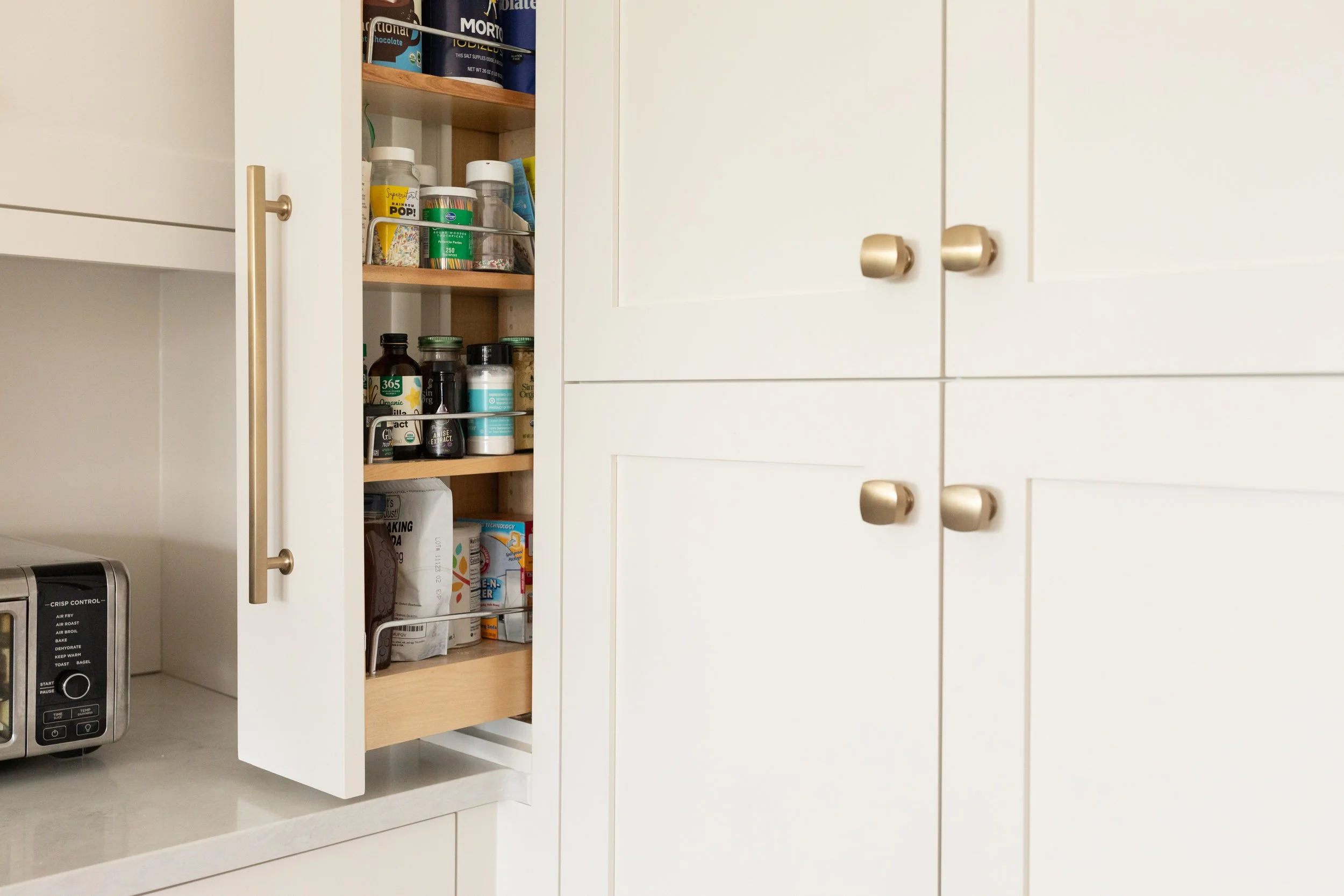 Open kitchen cabinet with stored vitamins, medicine bottles, and boxed food, next to a white kitchen door with gold handles, and a microwave on the countertop.