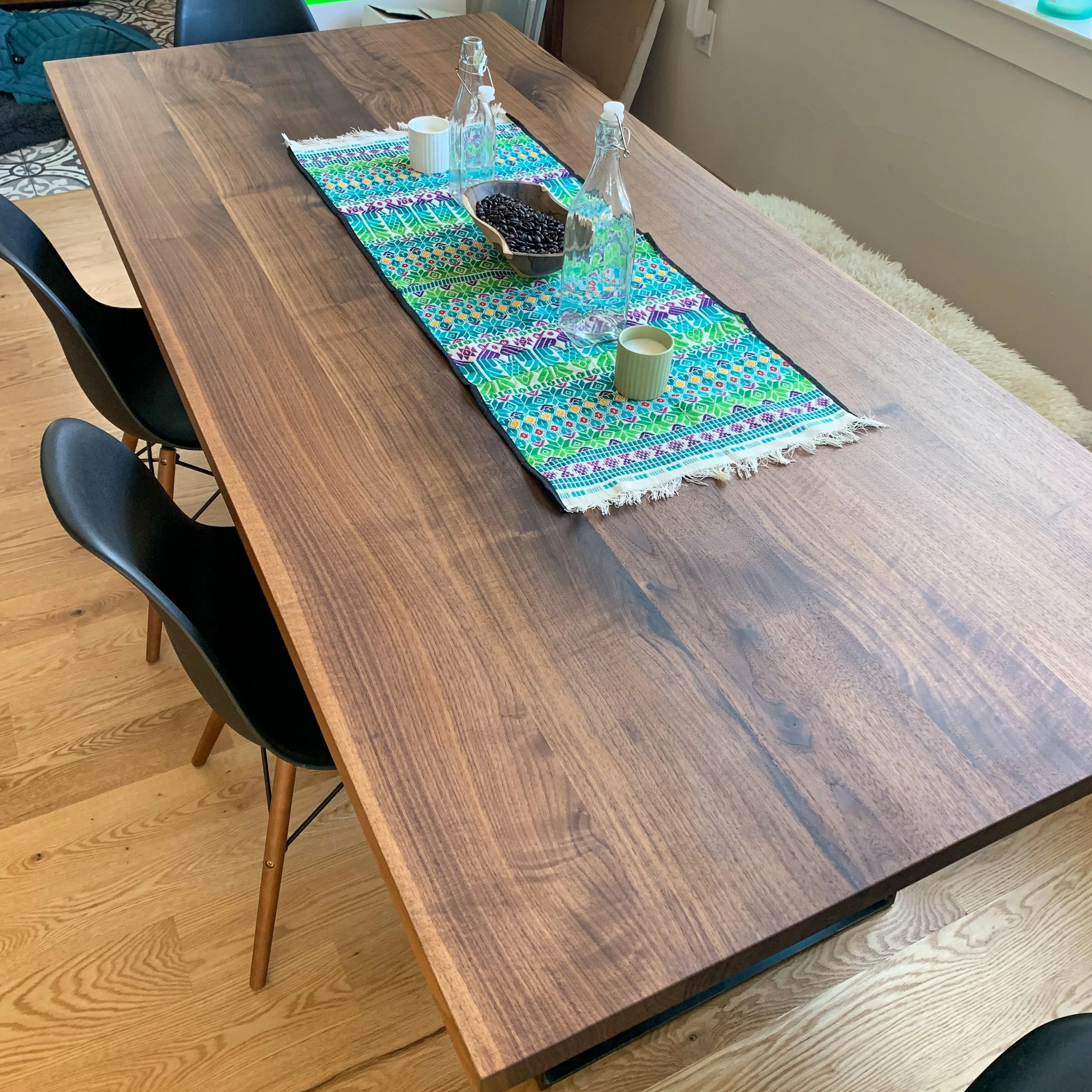 A wooden dining table with a colorful table runner, three glass bottles, two small cups, and a bowl of coffee beans.