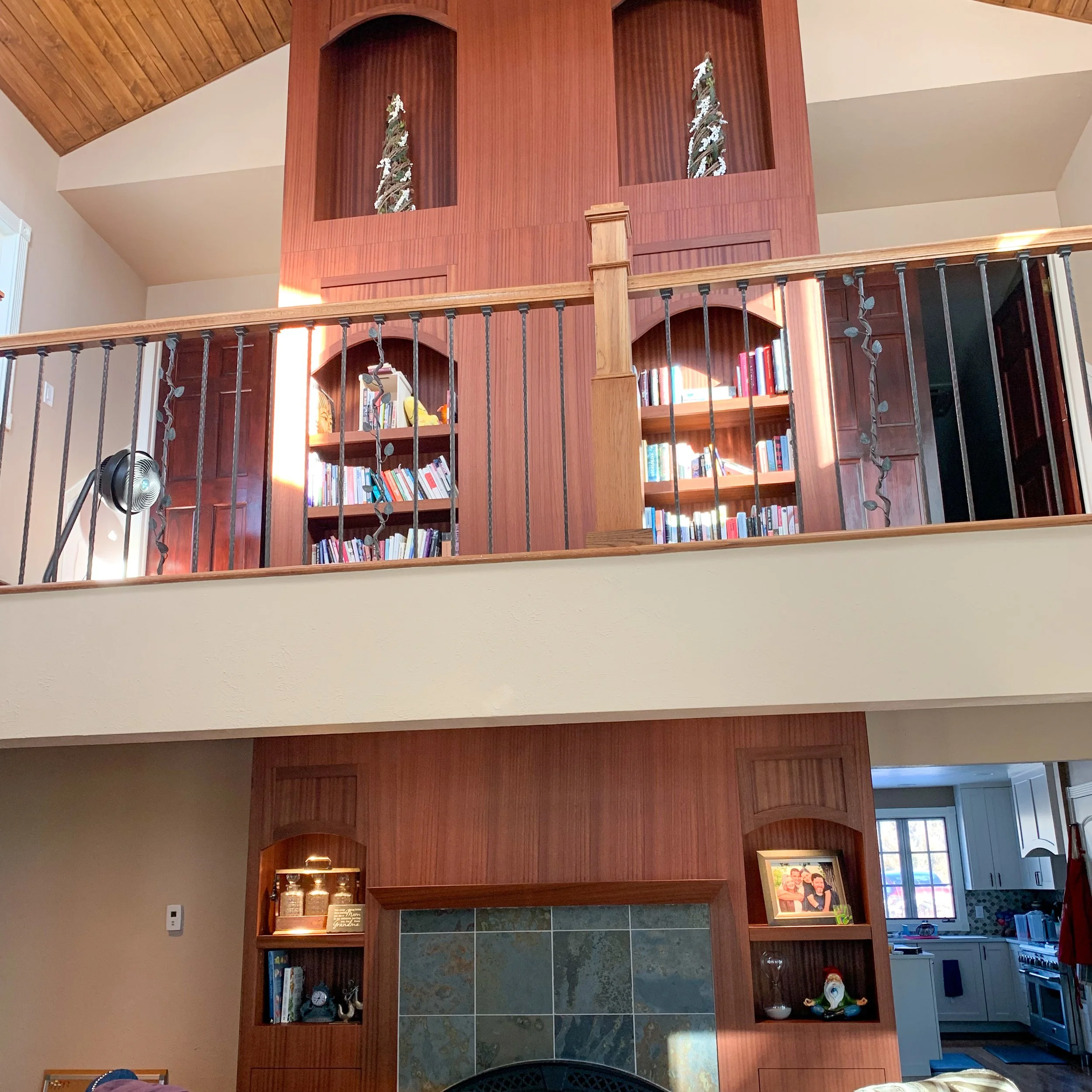 View of a wooden interior with a balcony railing, bookshelves filled with books, decorative items, and a partial view of a kitchen in the background.