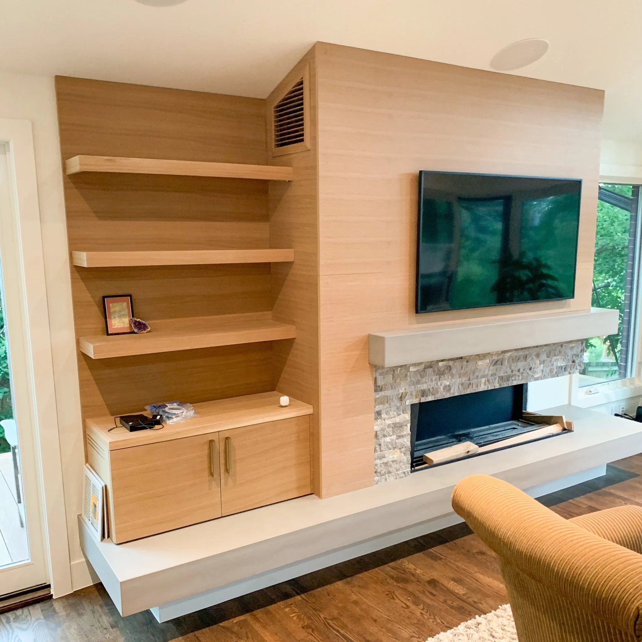 Living room corner with a wall-mounted flat-screen TV, a stone fireplace, a beige armchair, and wooden shelving units.