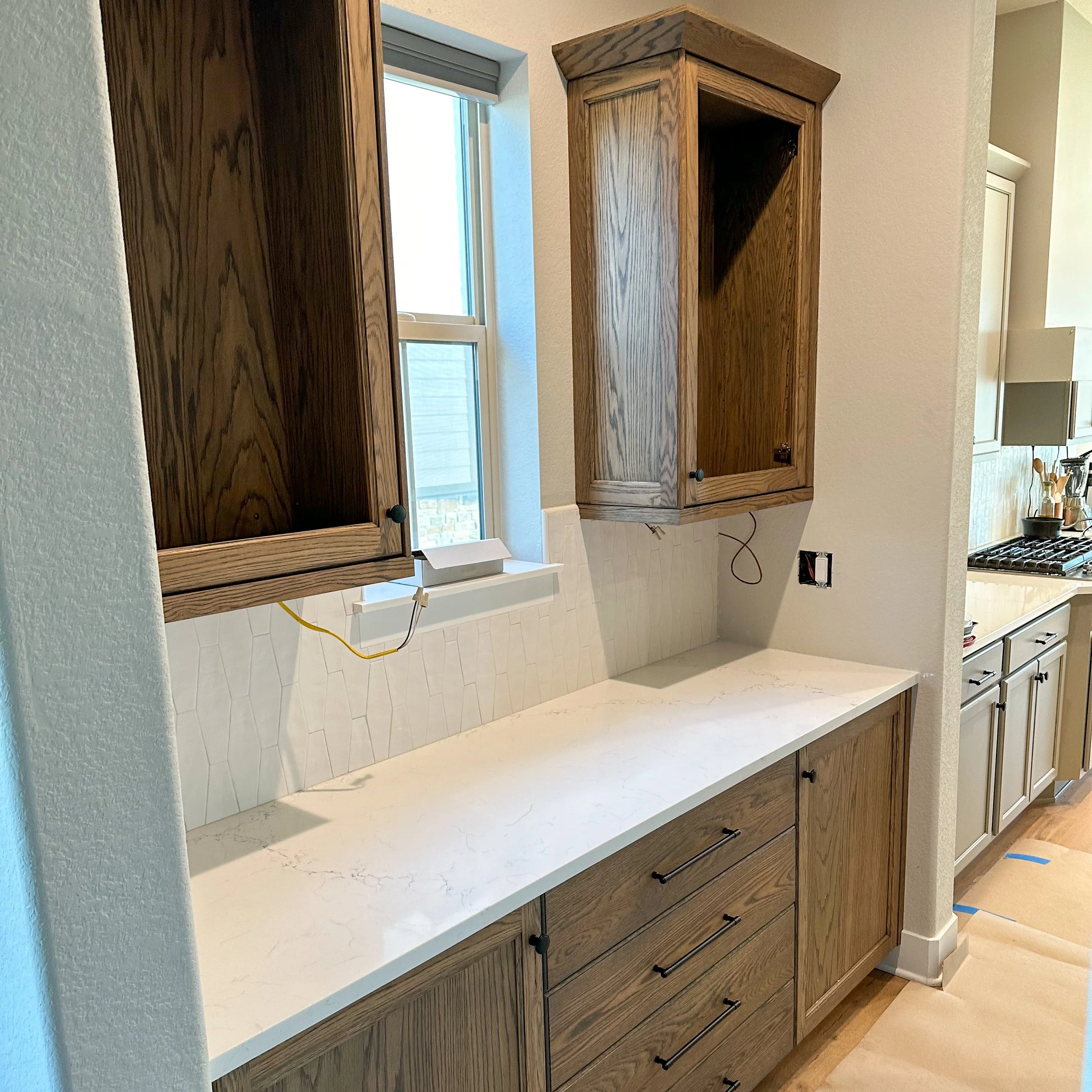Kitchen cabinet area with wooden upper and lower cabinets, a white marble countertop, a window, and an unfinished electrical outlet.