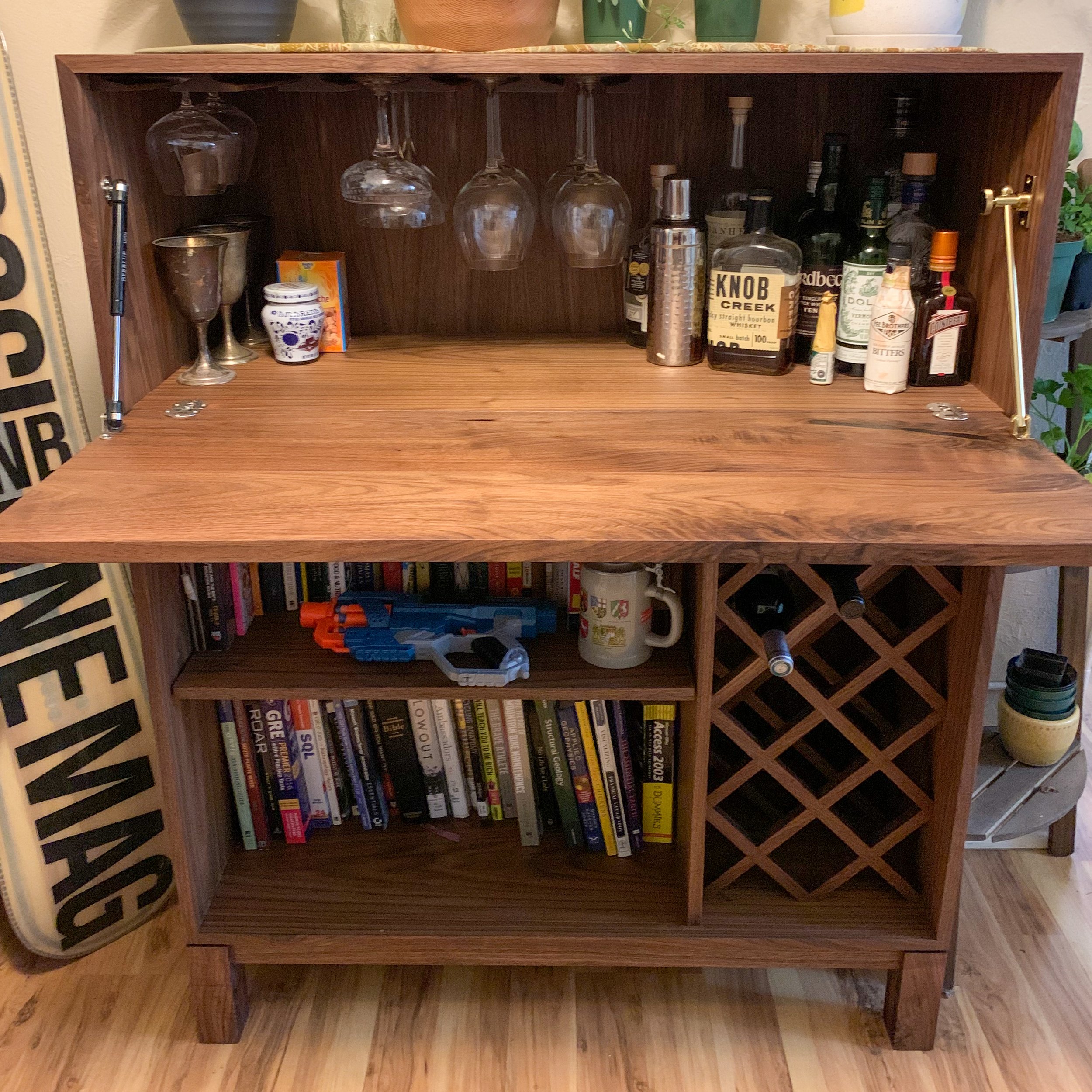 A wooden bar cabinet with a fold-down top, open shelf filled with books, compartments for wine bottles, and a collection of liquor bottles and glassware on top.