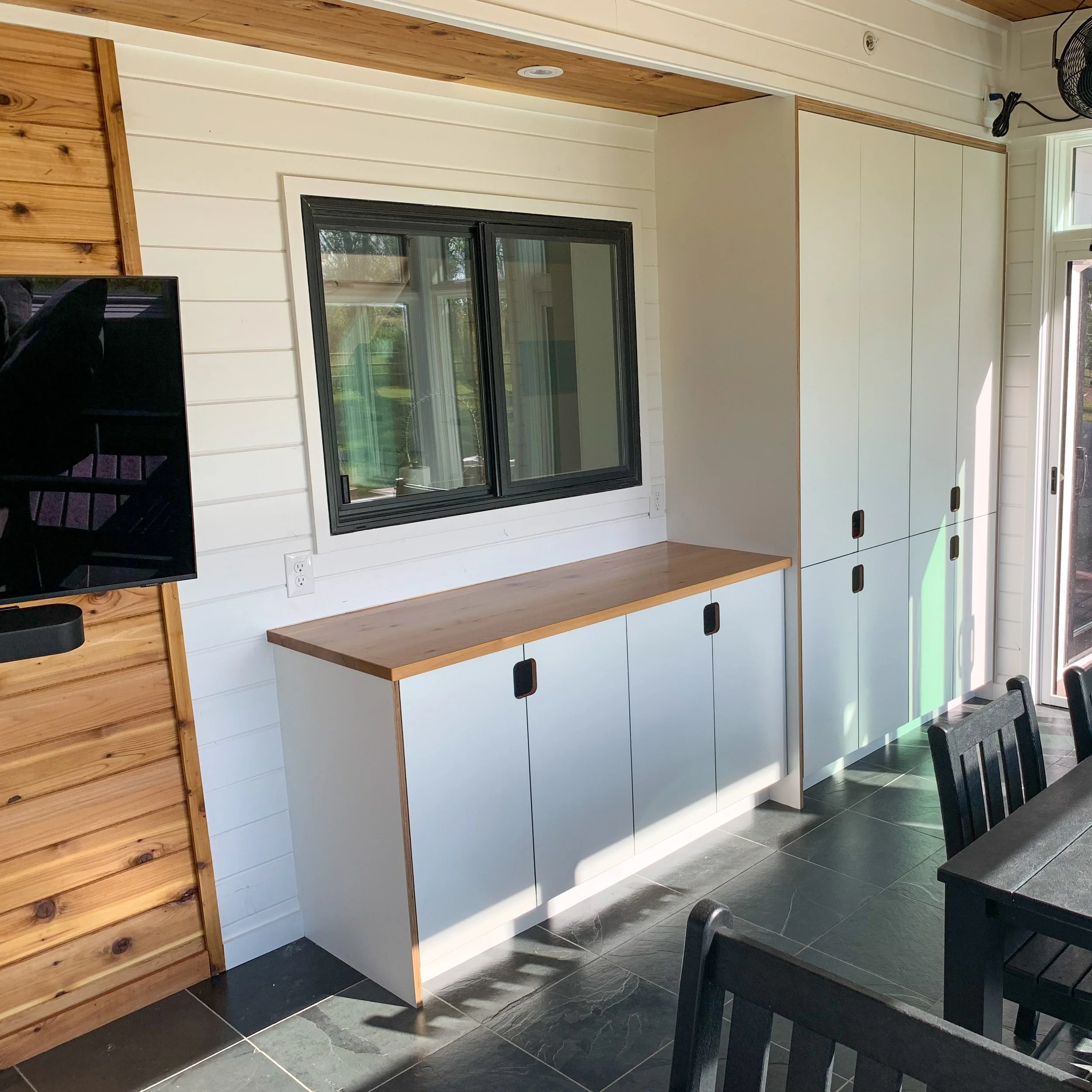 Interior of a modern room with black-framed window, white built-in cabinets with black handles, wooden countertop, black tiled floor, and a wall-mounted TV on a wood-paneled accent wall.