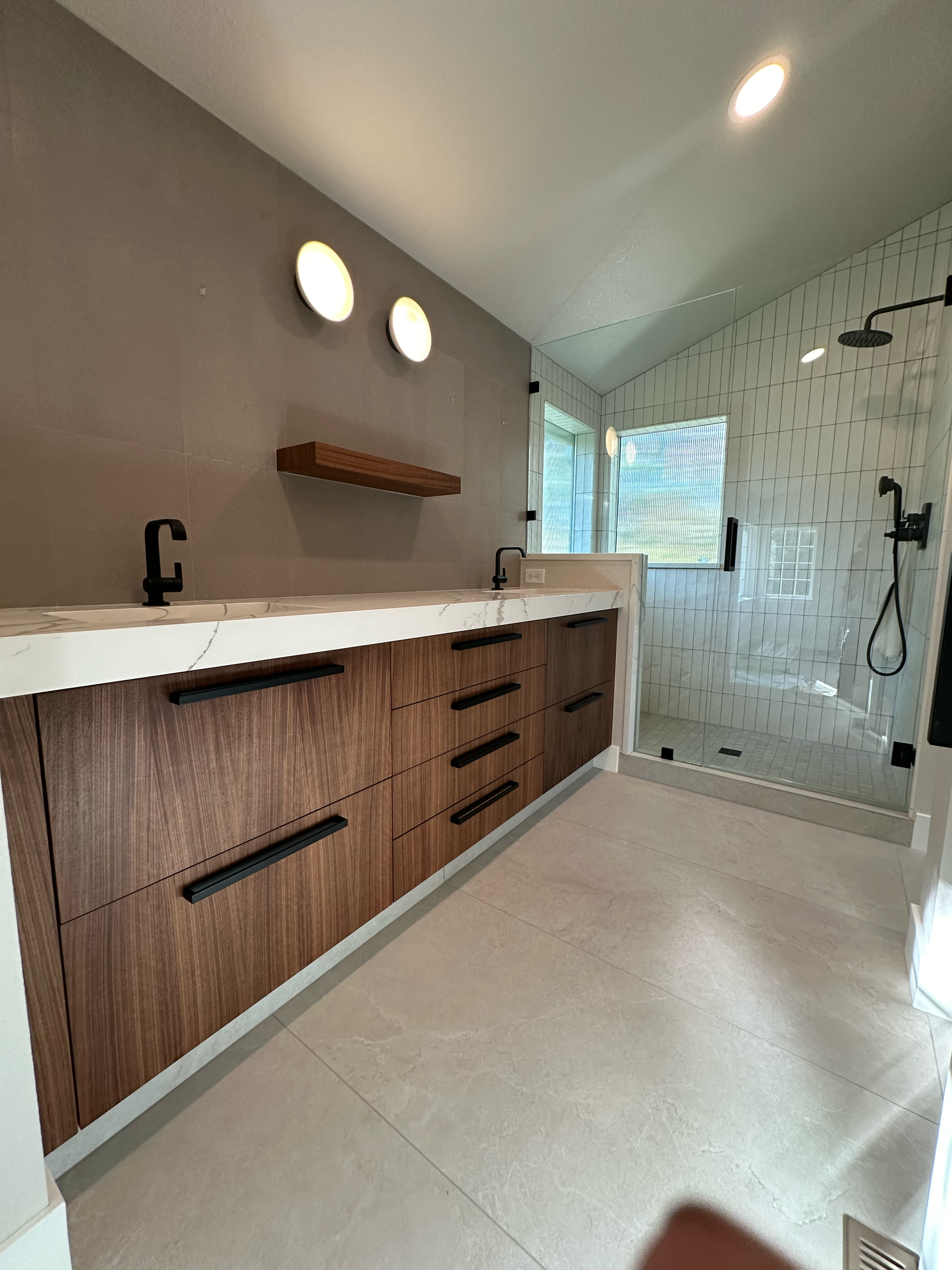 Modern bathroom with a shower area featuring glass doors, black fixtures, and a tile wall, and a vanity with wood drawers and black handles, marble countertop, beige floor tiles, and a small wooden shelf unit.