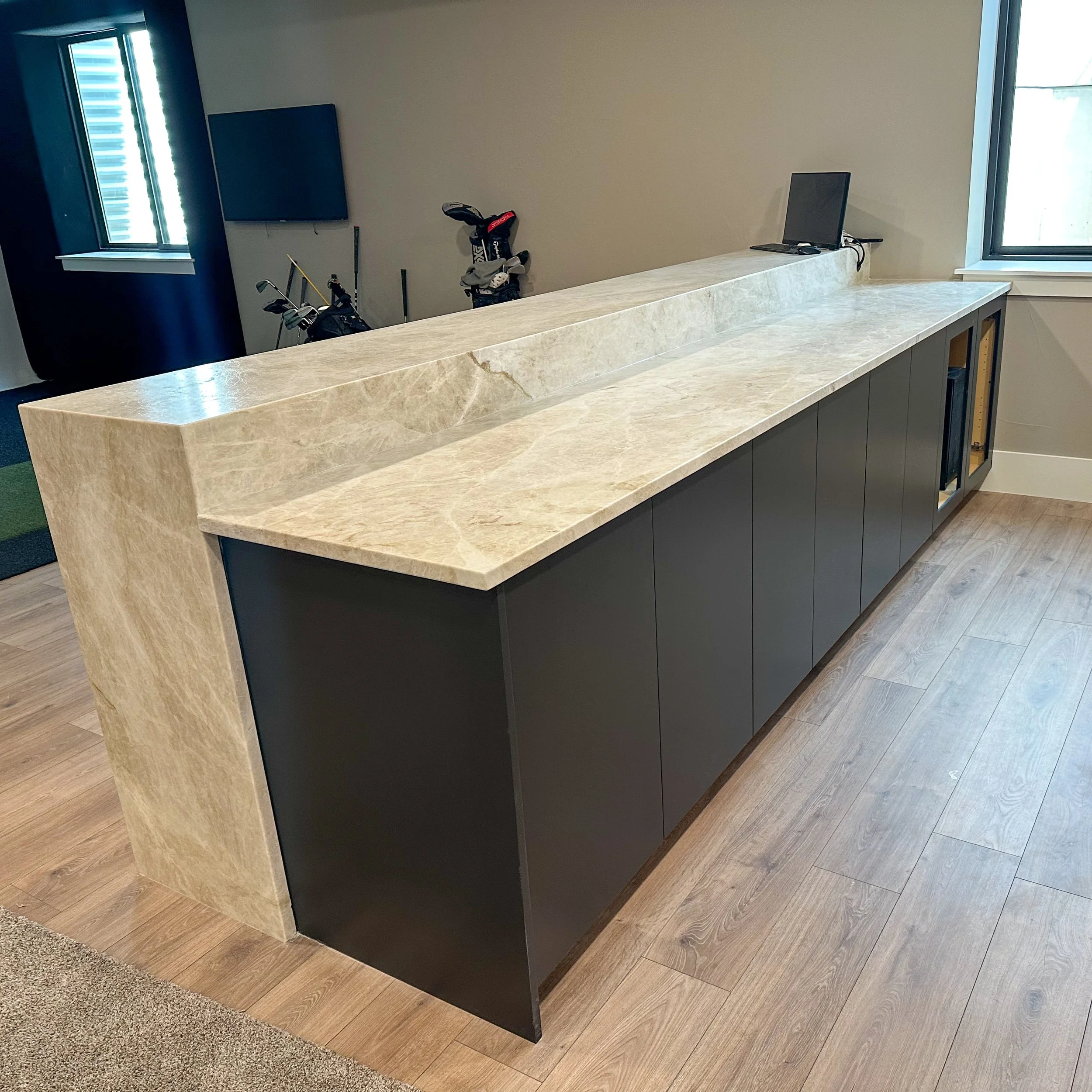 A modern reception desk with a beige marble top and black base, located in an indoor space with wooden flooring and two windows.