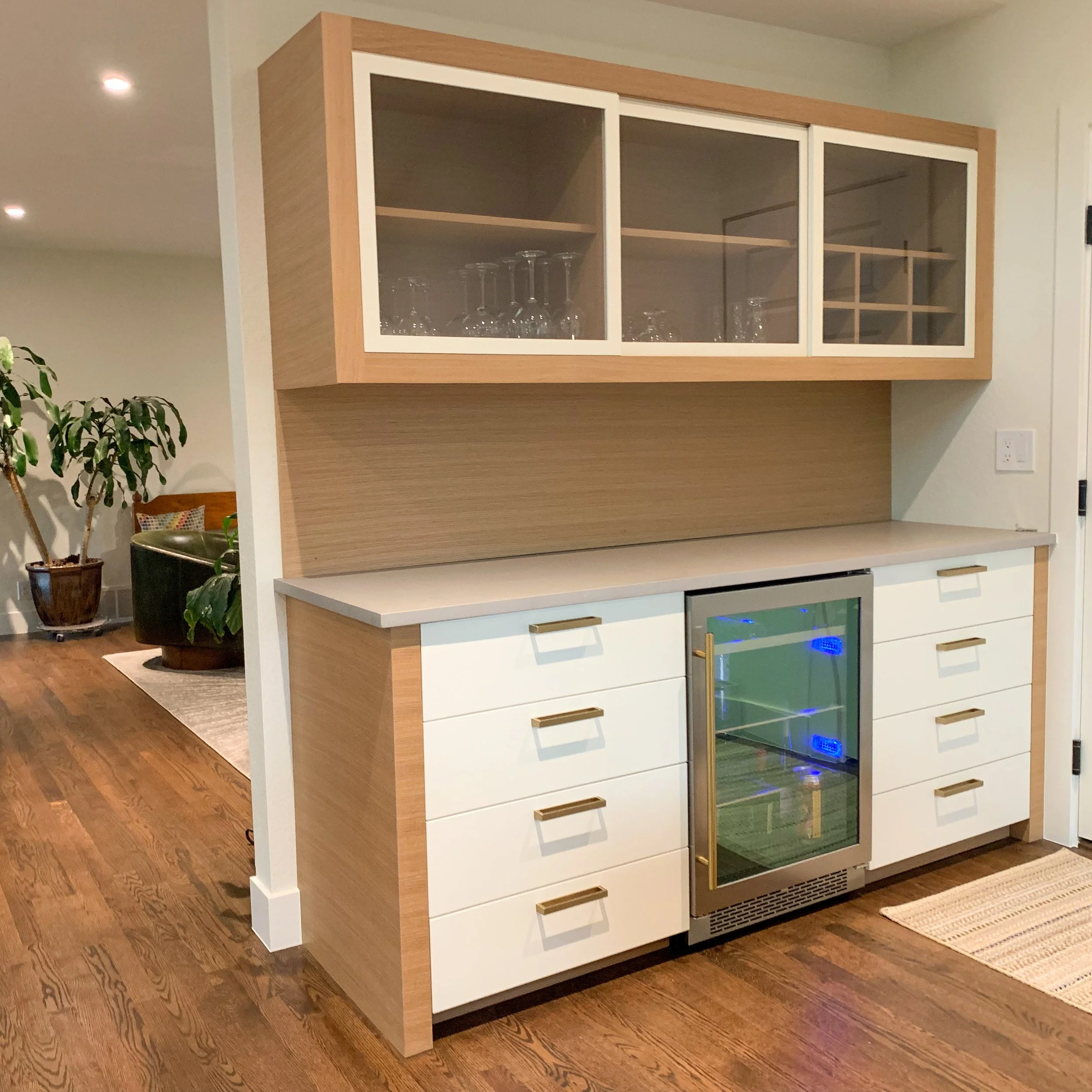 Modern kitchen bar area with white cabinets, gold handles, a built-in mini fridge with a glass door, and upper glass-fronted cabinets containing wine glasses. Wooden flooring and a beige rug are visible.