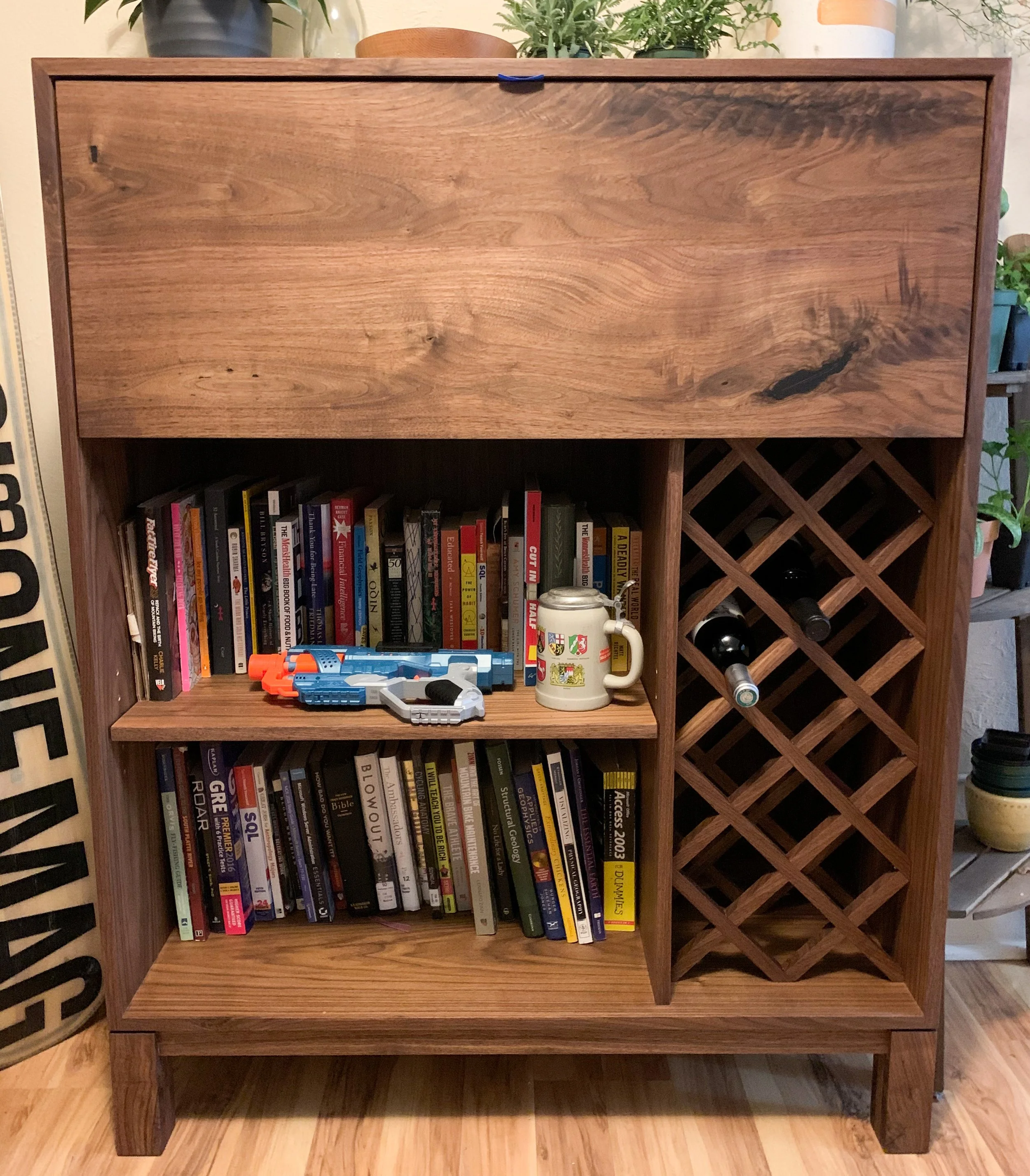 A wooden bookshelf containing books, a toy gun, and a beer stein, with wine bottle holders on the right side and potted plants on top.