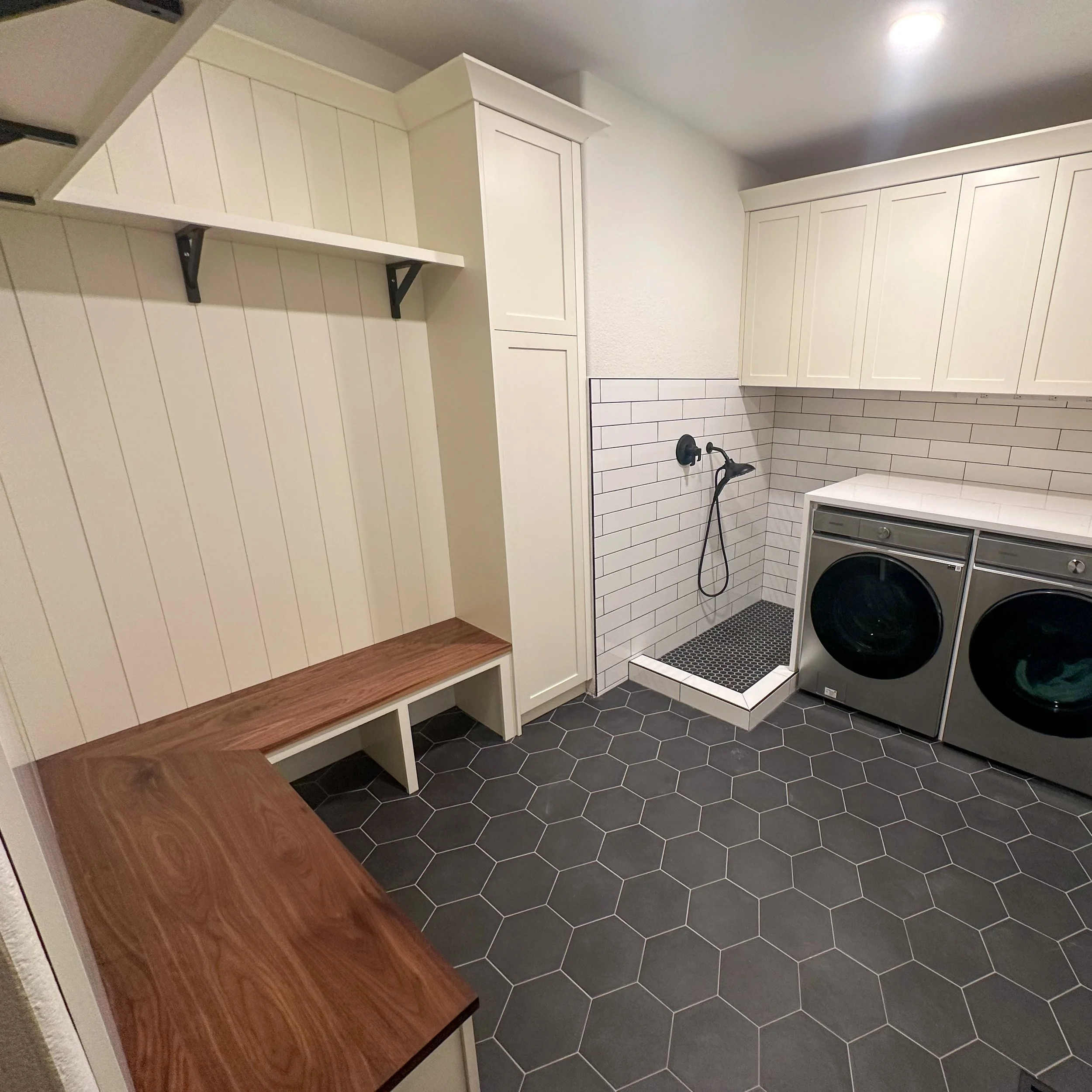 Laundry room with white cabinets, a wooden bench, black hexagonal floor tiles, a washing machine and dryer, and a black and white tiled shower area.