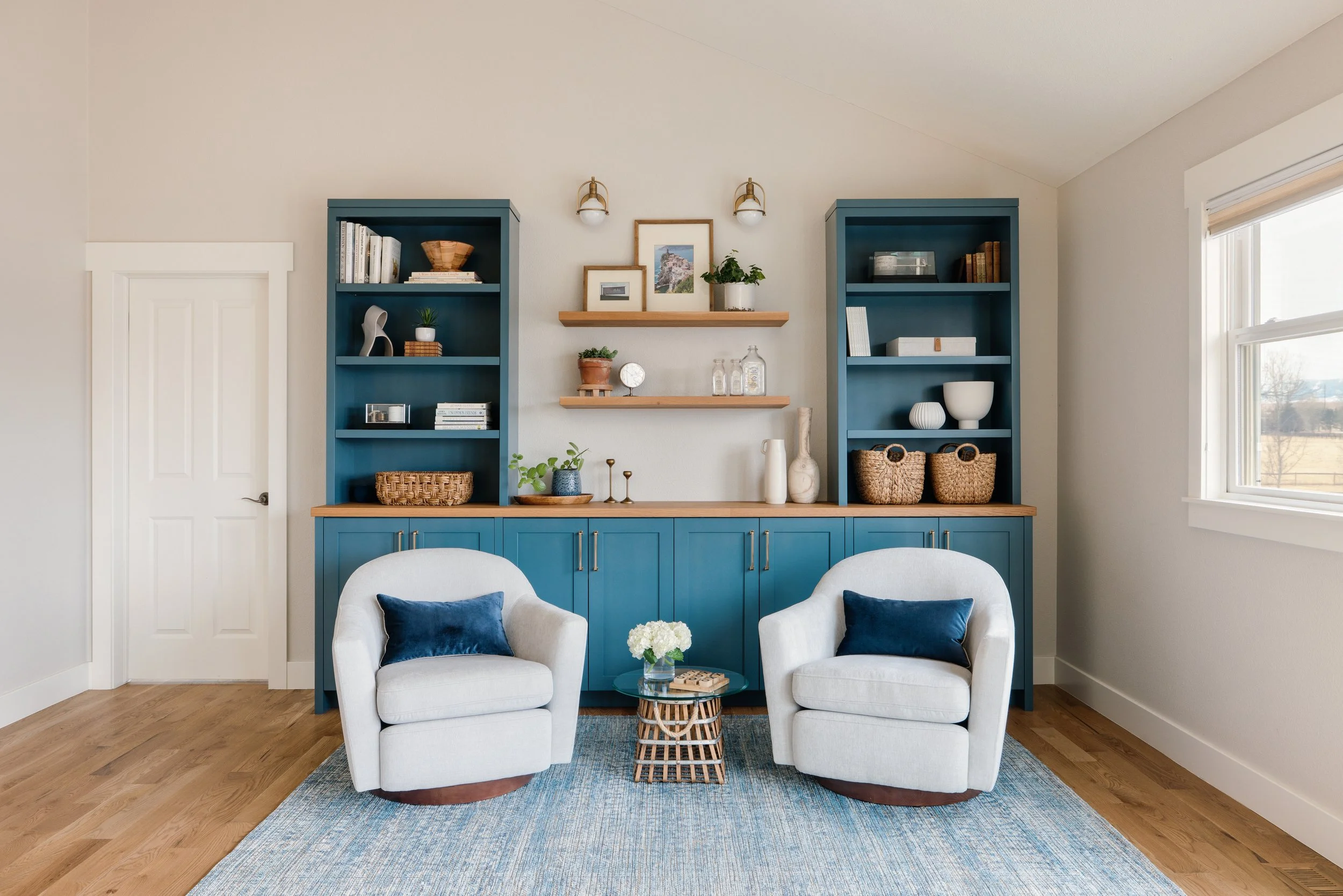 Living room with two white armchairs, blue pillows, a small wicker side table with a glass top and a floral arrangement, and a blue built-in bookshelf with open shelves and closed cabinets, decorated with books, baskets, vases, and framed photos. A w