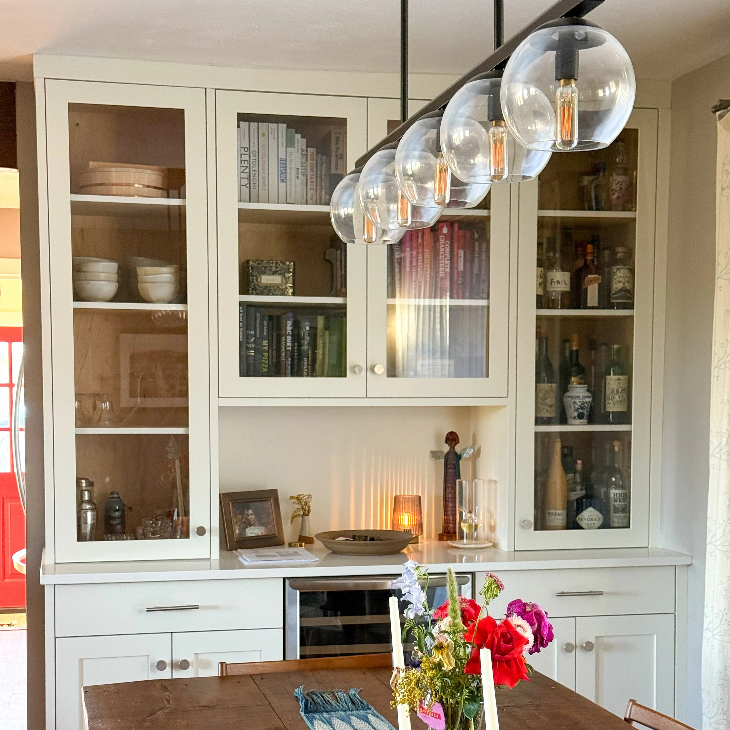 A white kitchen cabinet with glass doors displaying books, bowls, and bottles, with a wooden table in foreground holding a vase of colorful flowers.