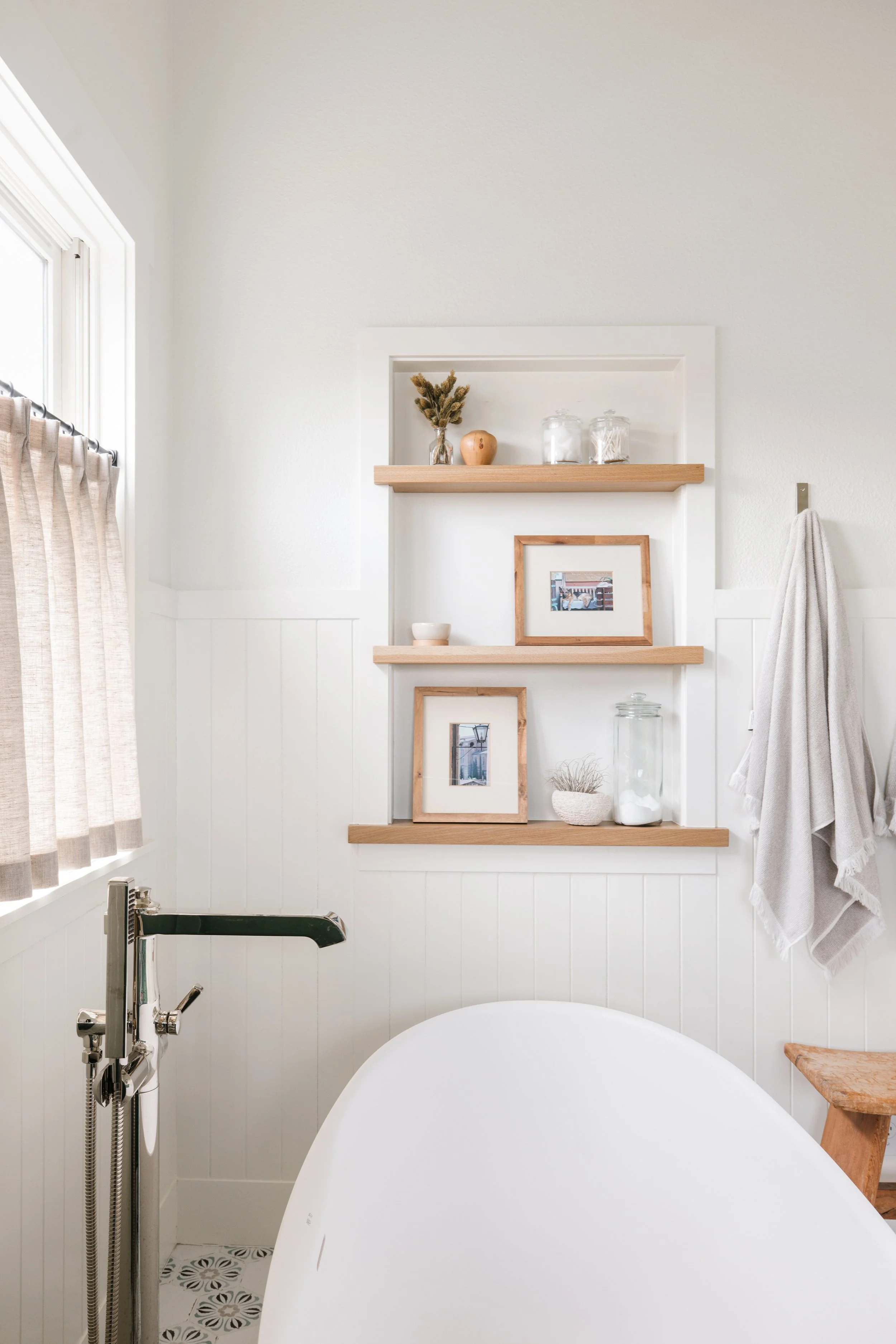 Cozy bathroom with a white bathtub, wood shelves with framed pictures, jars, a small bowl, and decorative items, beige curtains on a window, and a white wall with a towel hanging.