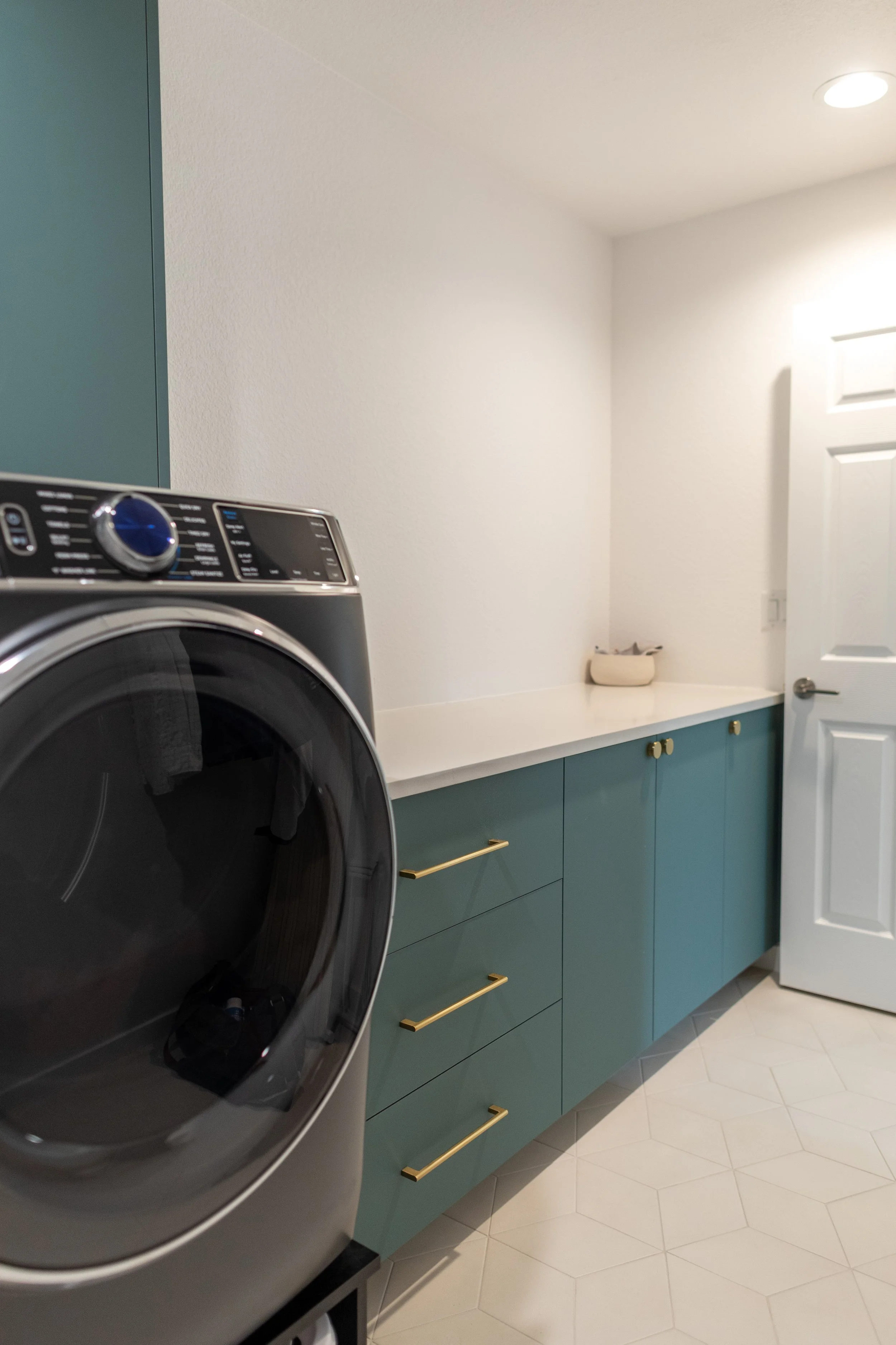 A laundry room with a front-loading washing machine, teal cabinets with gold handles, a white countertop, and a white door.