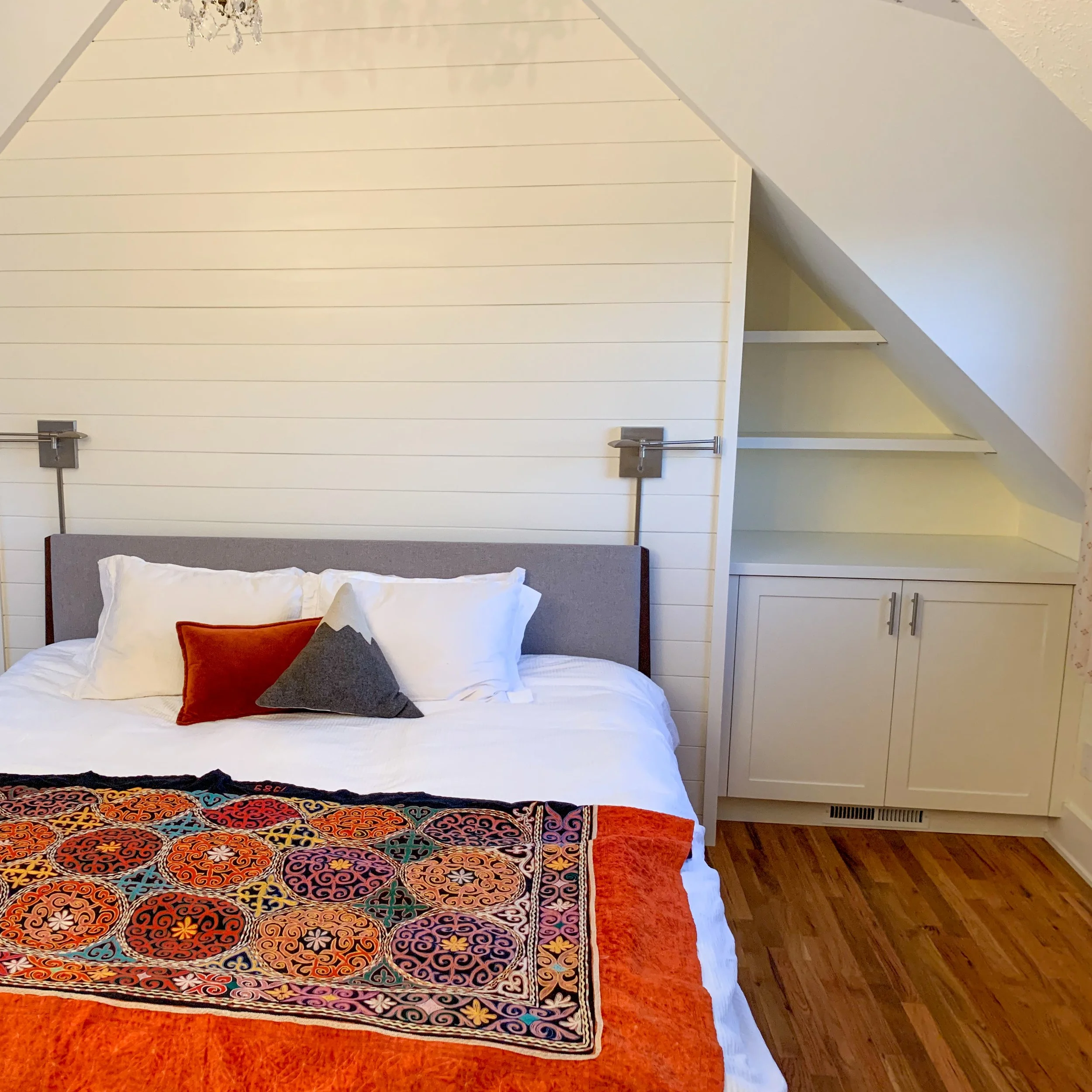 A bedroom with a bed, decorative pillows, a colorful quilt, a white dresser, and built-in shelves under a sloped ceiling.