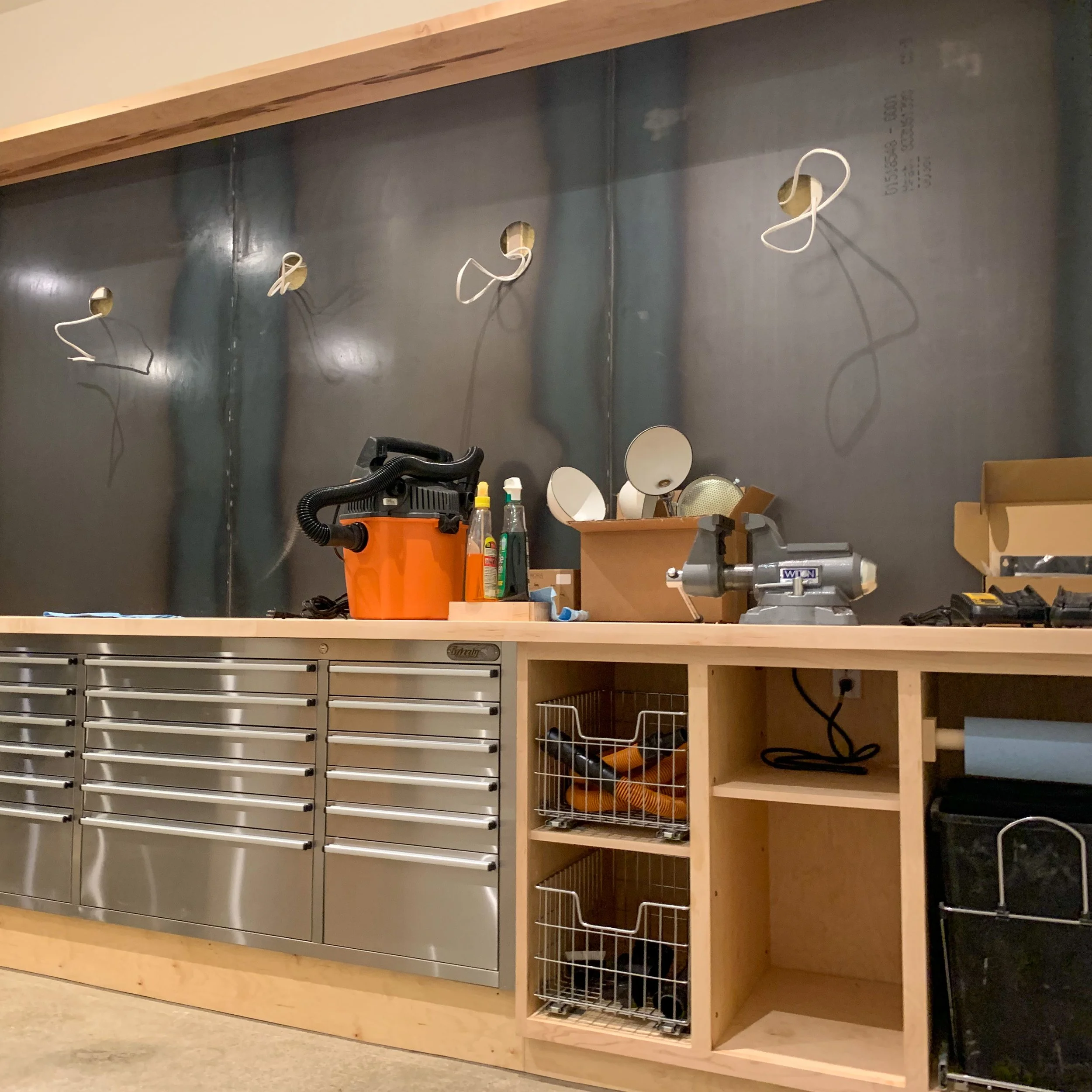 Workshop with metal cabinet and wooden workbench, tools, and supplies, wall with electrical outlets and hanging wires.