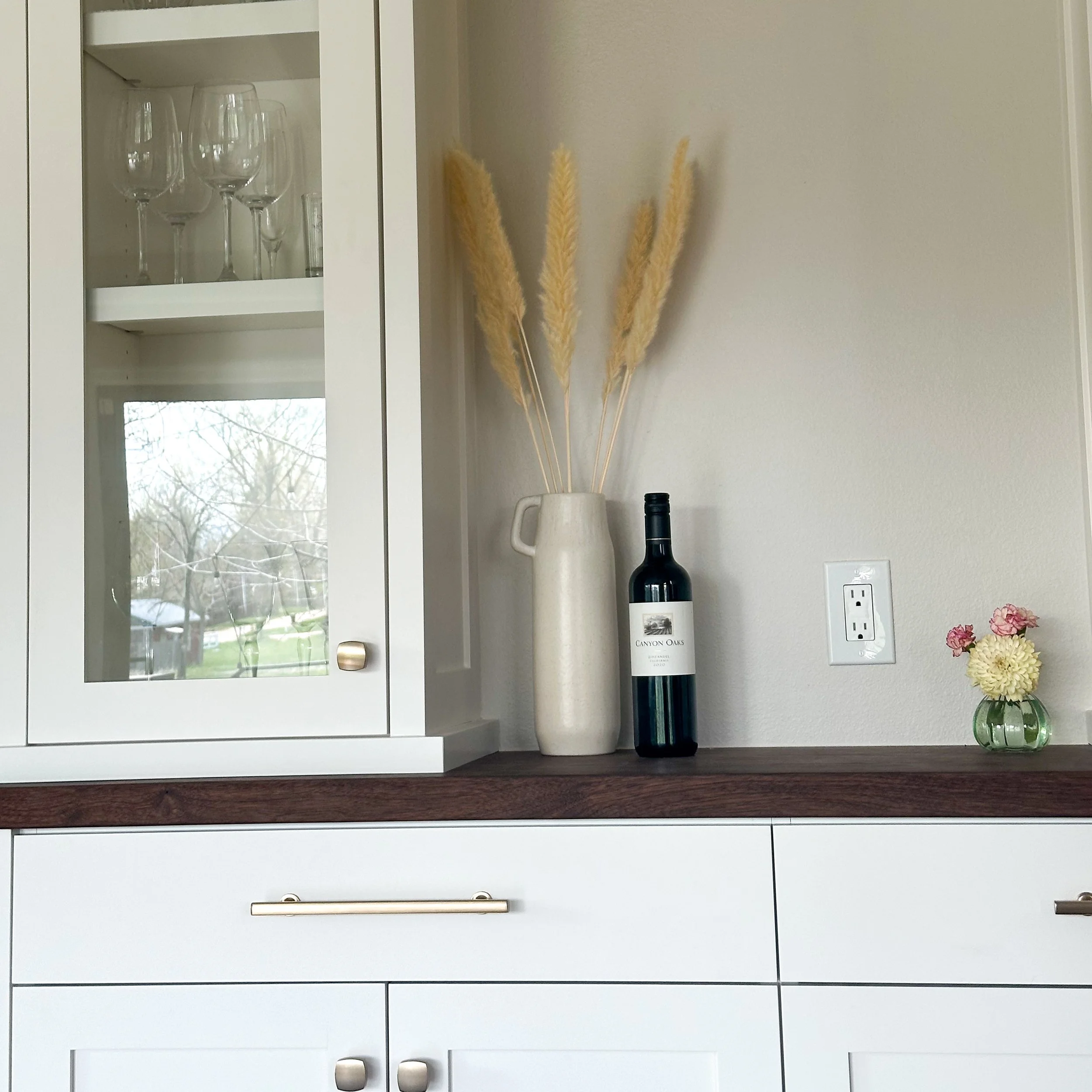 White cabinet with a glass door displaying wine glasses, a countertop with a vase of dried grass, a bottle of wine, a small glass vase with pink and yellow flowers, and a power outlet on a beige wall.