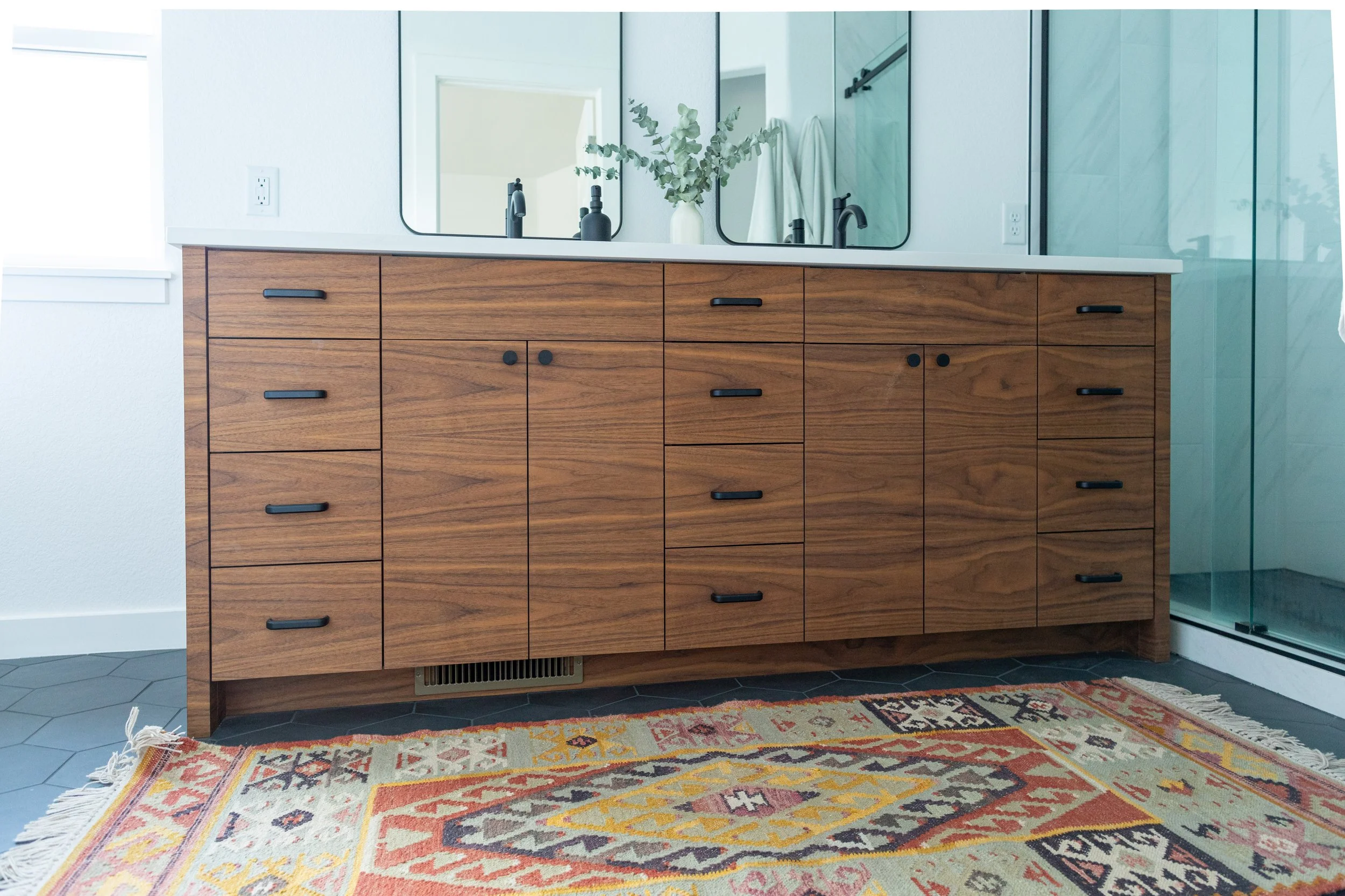 A wooden vanity with multiple drawers and black handles, topped with a white countertop. Behind the vanity are three mirrors, a vase with greenery, and a black soap dispenser. To the right is a glass shower door. The floor has dark tiles, and a decor