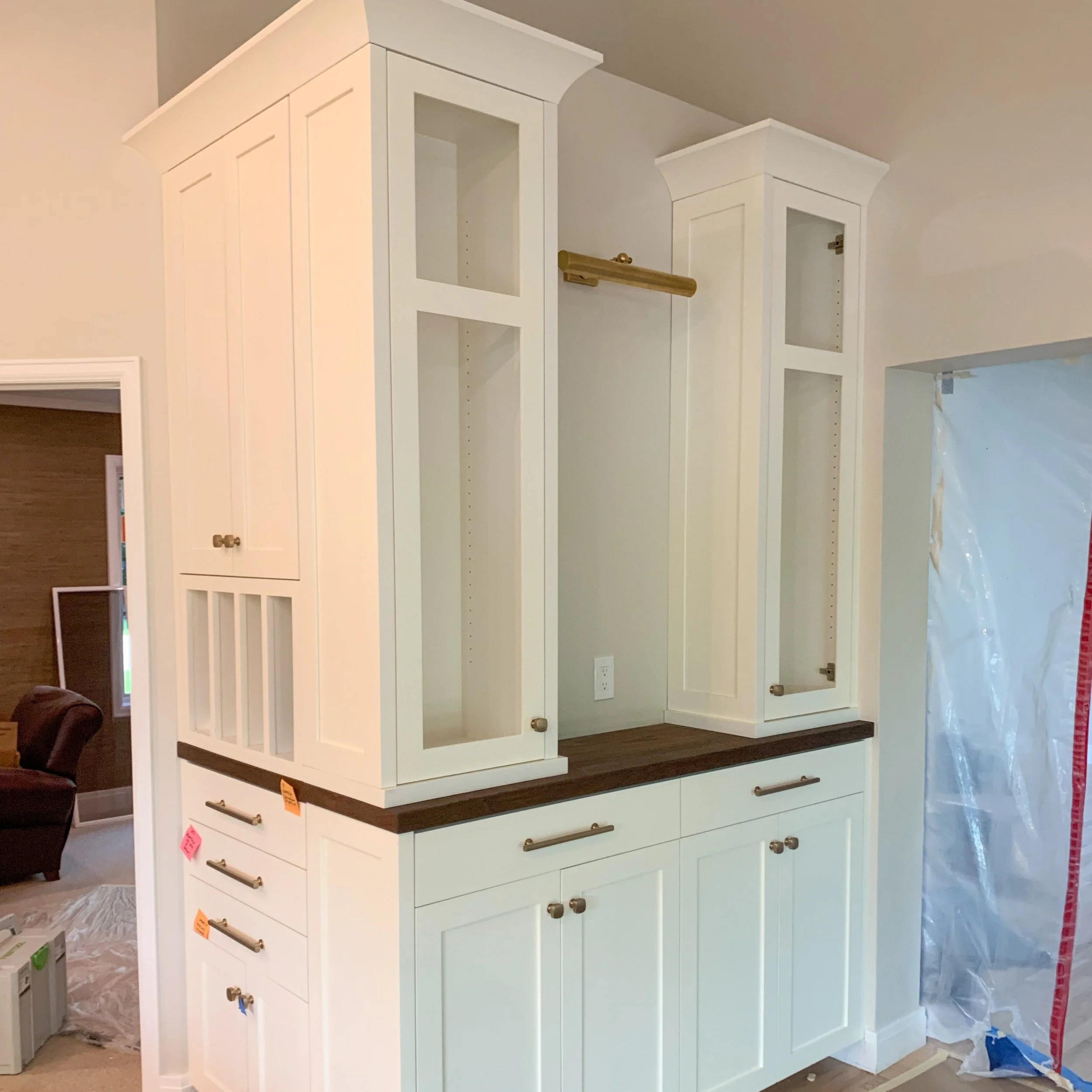White kitchen cabinets with a dark wood countertop, some drawers with handles, and open glass-front cabinets. There is a white electrical outlet in the center, and the area is under construction or renovation.
