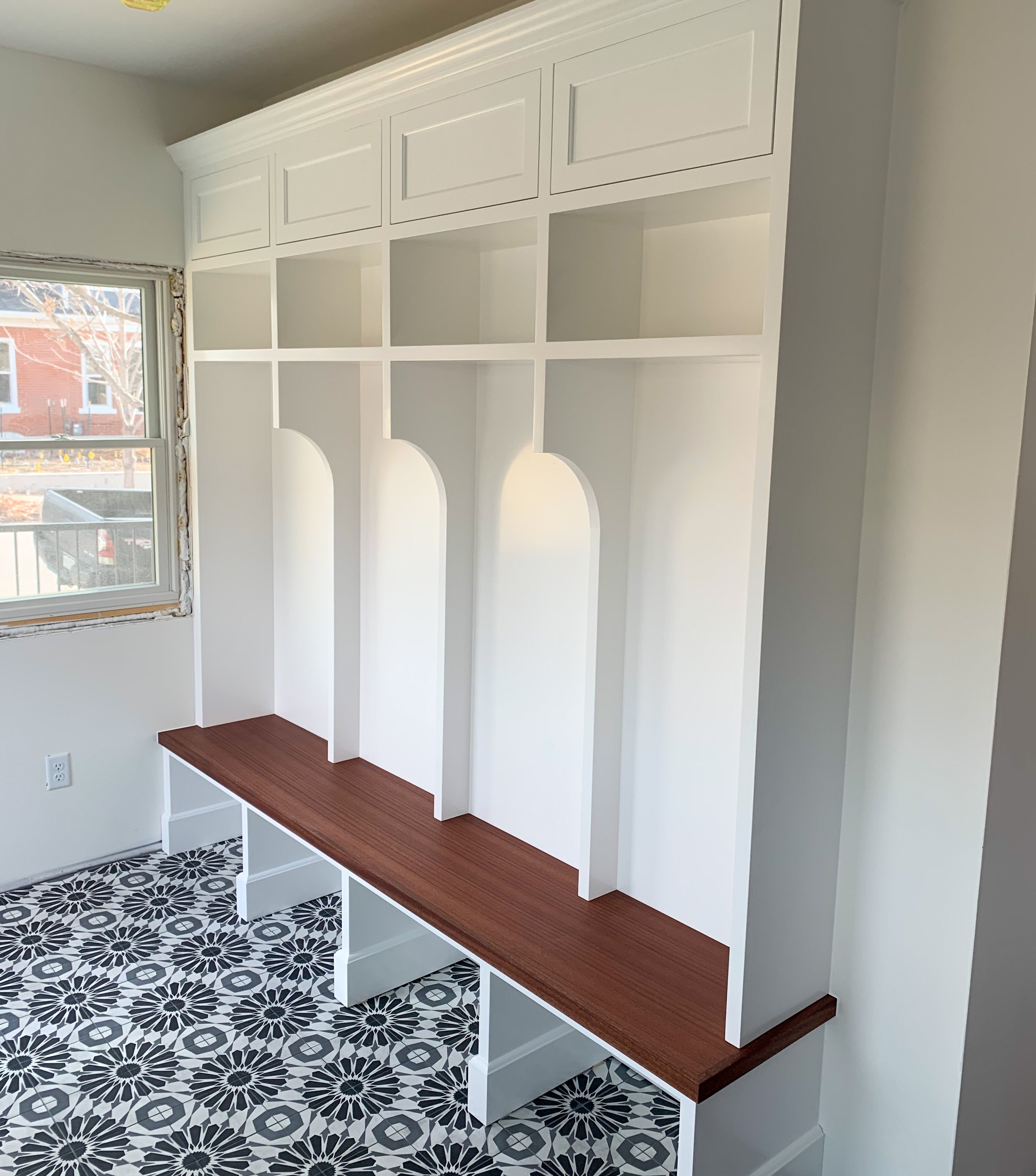 Empty built-in white wooden shelving unit with a brown wooden countertop, set against a white wall near a window in a room with black and white patterned floor tiles.