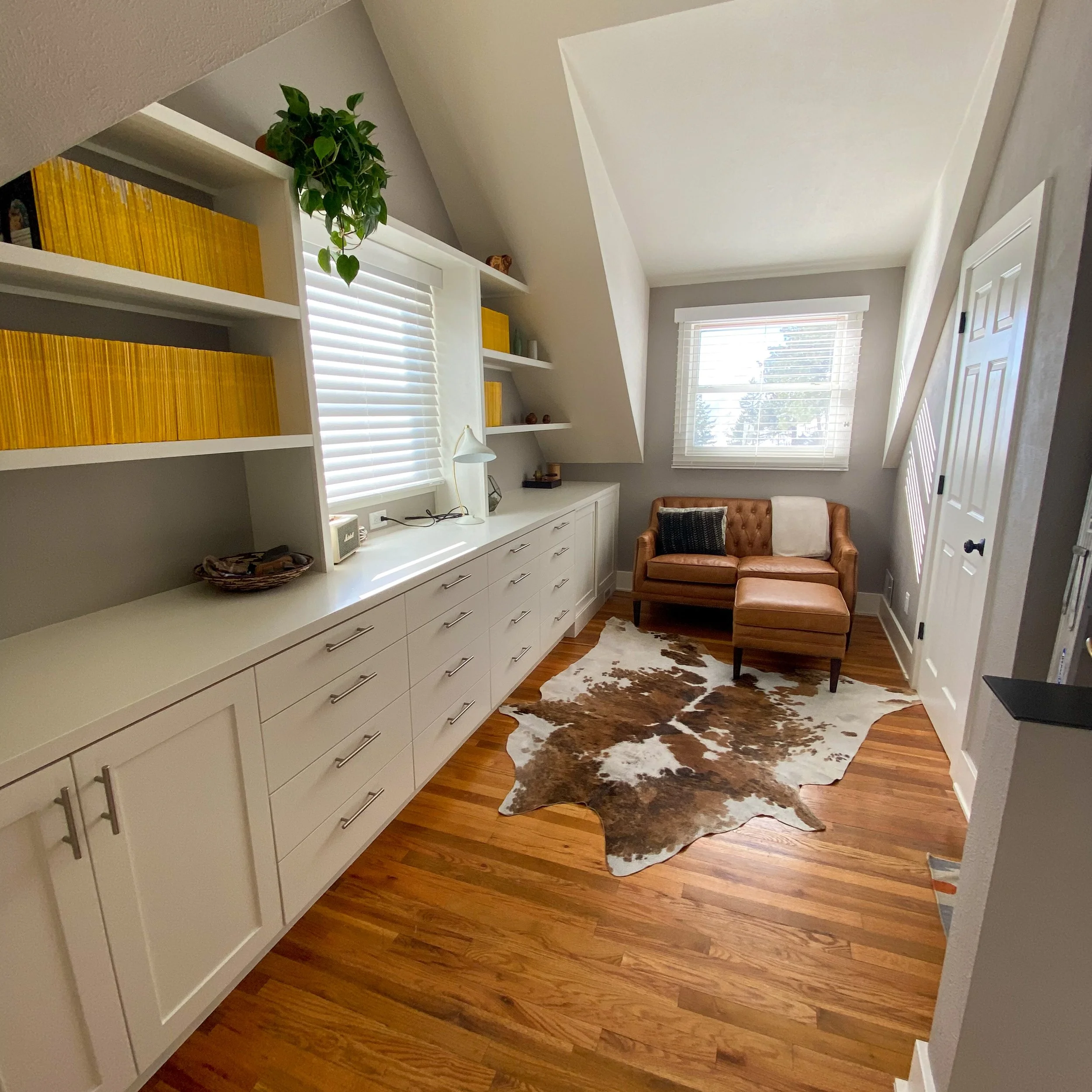 A cozy room with white built-in cabinetry along the left wall, topped with bookshelves filled with yellow books and small decor items. The room features two windows with white blinds, a tan leather sectional sofa with a black pillow and white blanket