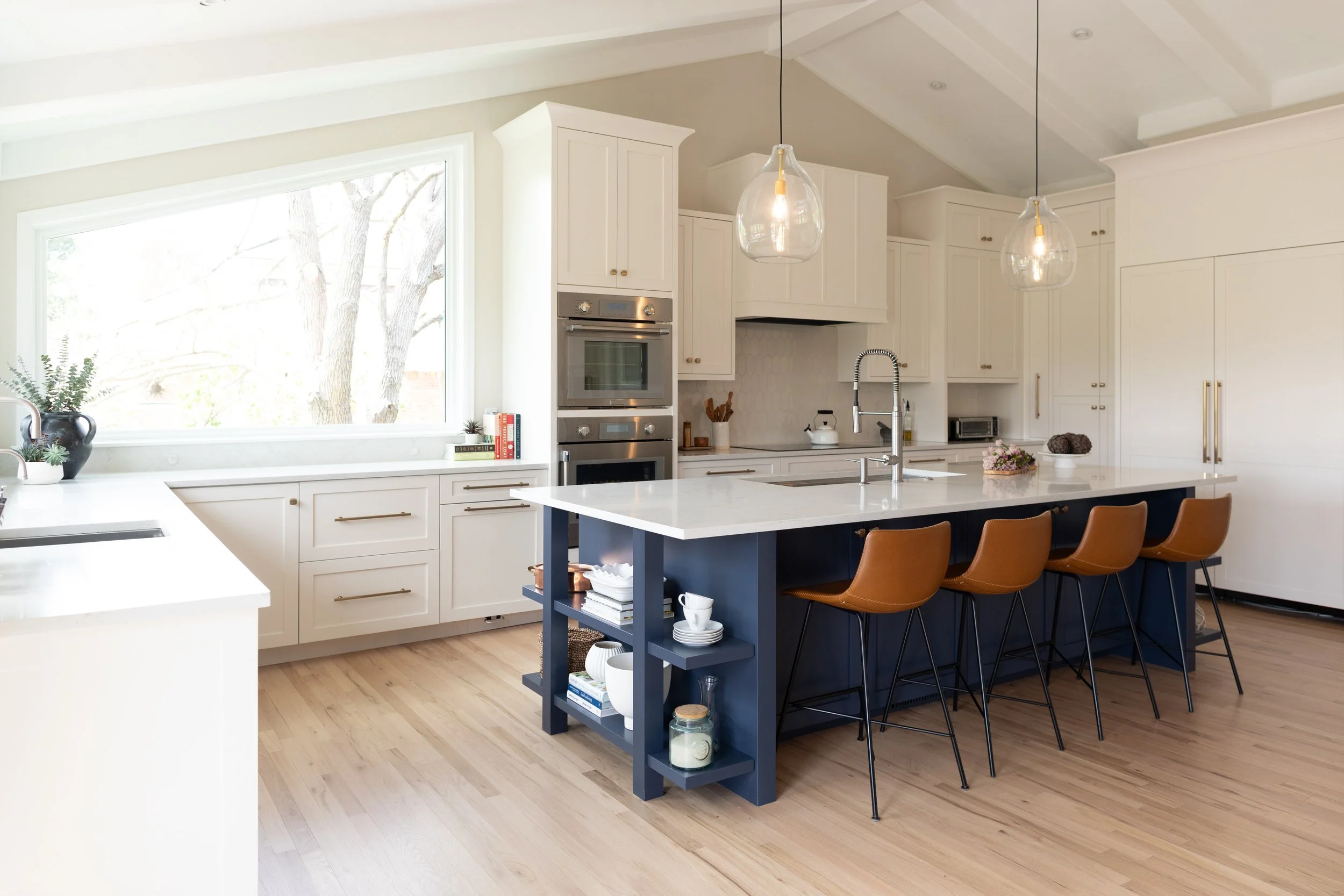 Modern kitchen with white cabinets, marble countertops, and a large window. There are four brown chairs at a navy blue island with open shelves, pendant lights hanging above, and stainless steel appliances.
