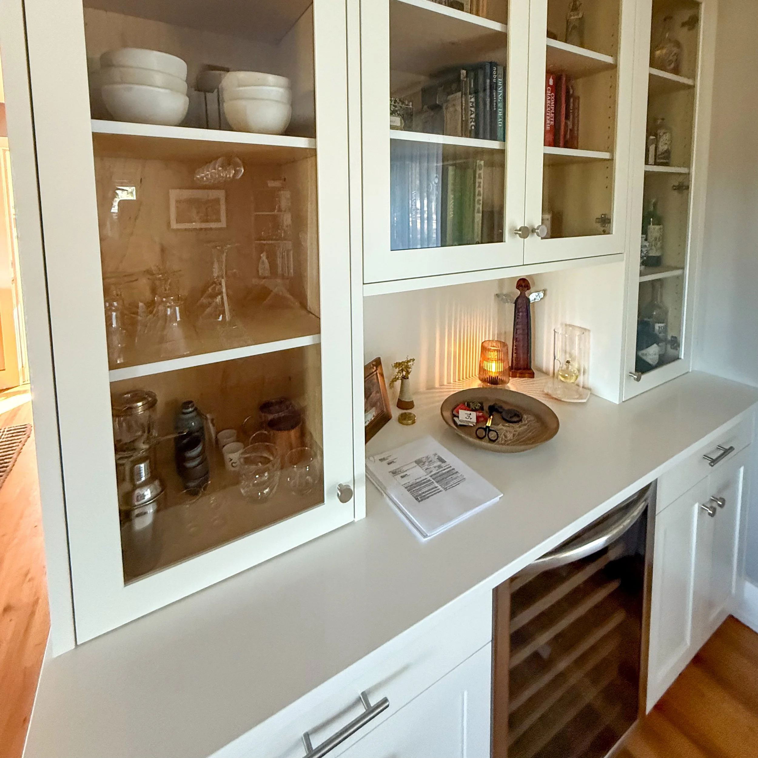 A white kitchen cabinet with glass doors displaying dishes, glasses, and books, a countertop with a tray, scissors, and documents, and a wine cooler below.