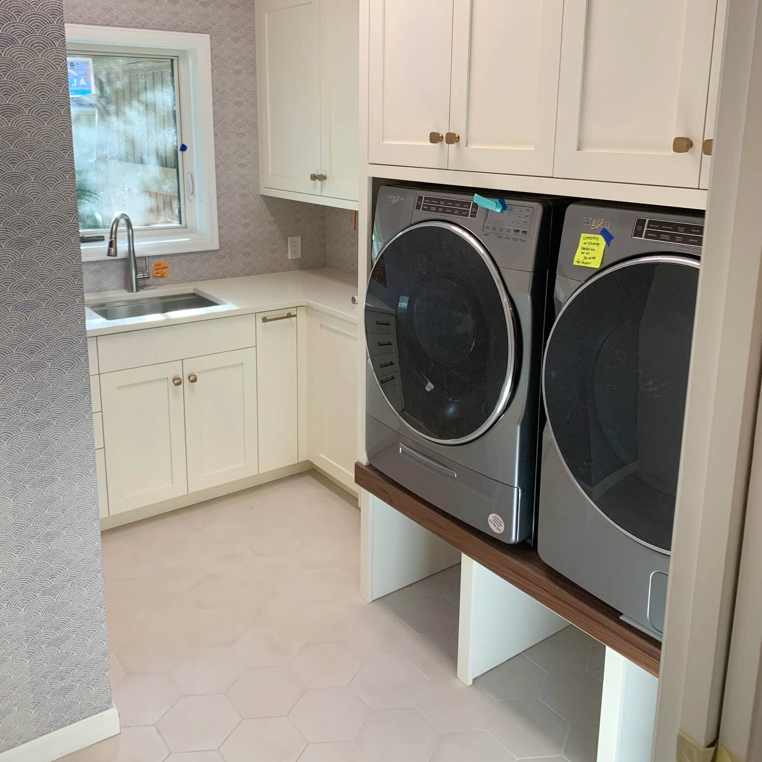 Laundry room with white cabinets, a small stainless steel sink, a window, and a front-loading washer and dryer stacked on a wooden shelf. The walls have wallpaper with a scallop pattern, and the floor has hexagonal tiles.