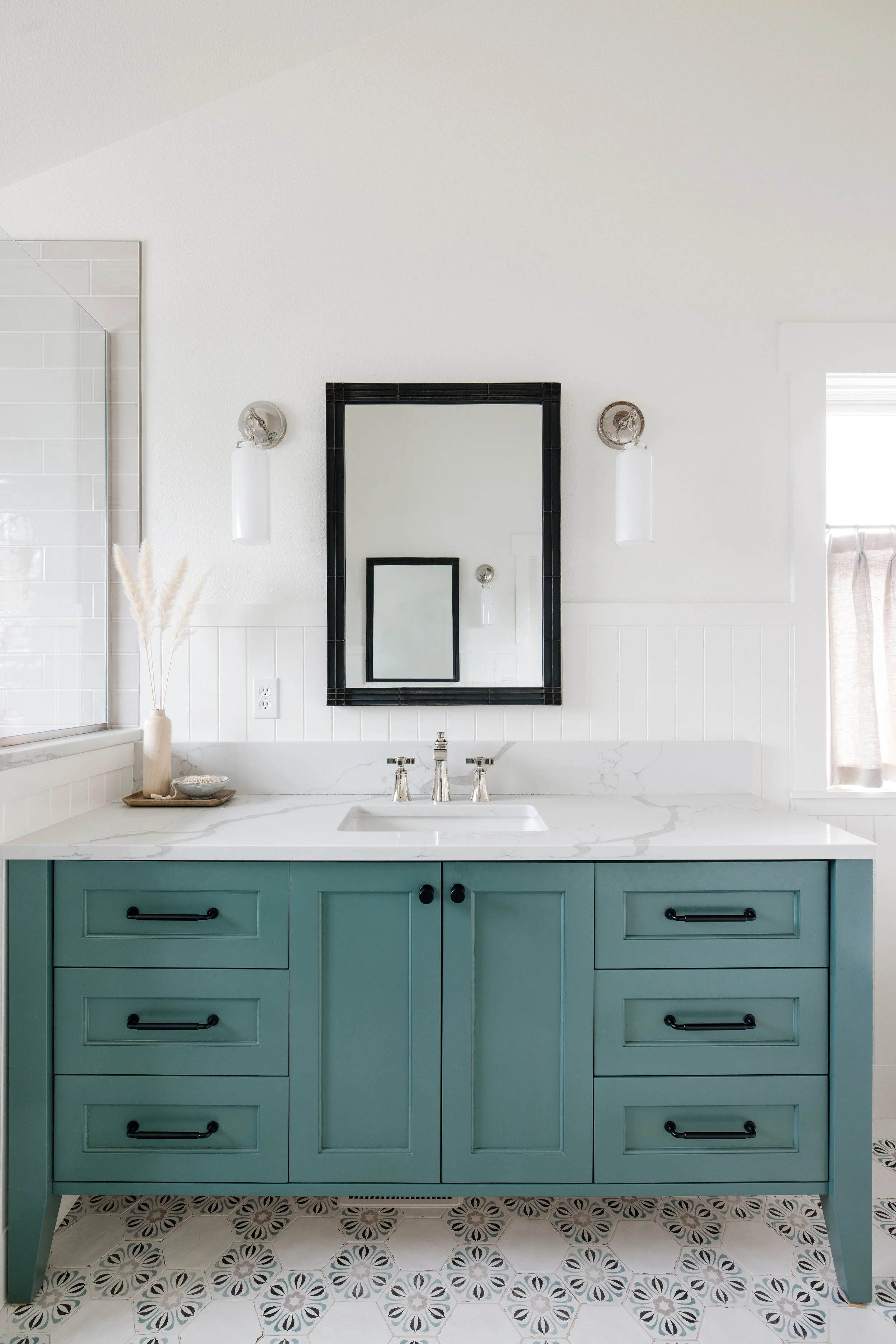 A bathroom vanity with teal cabinets, white marble countertop, two drawer handles, and a sink with a faucet, with a black-framed mirror mounted on the wall above, flanked by two wall fixtures, and decorative items on the countertop.