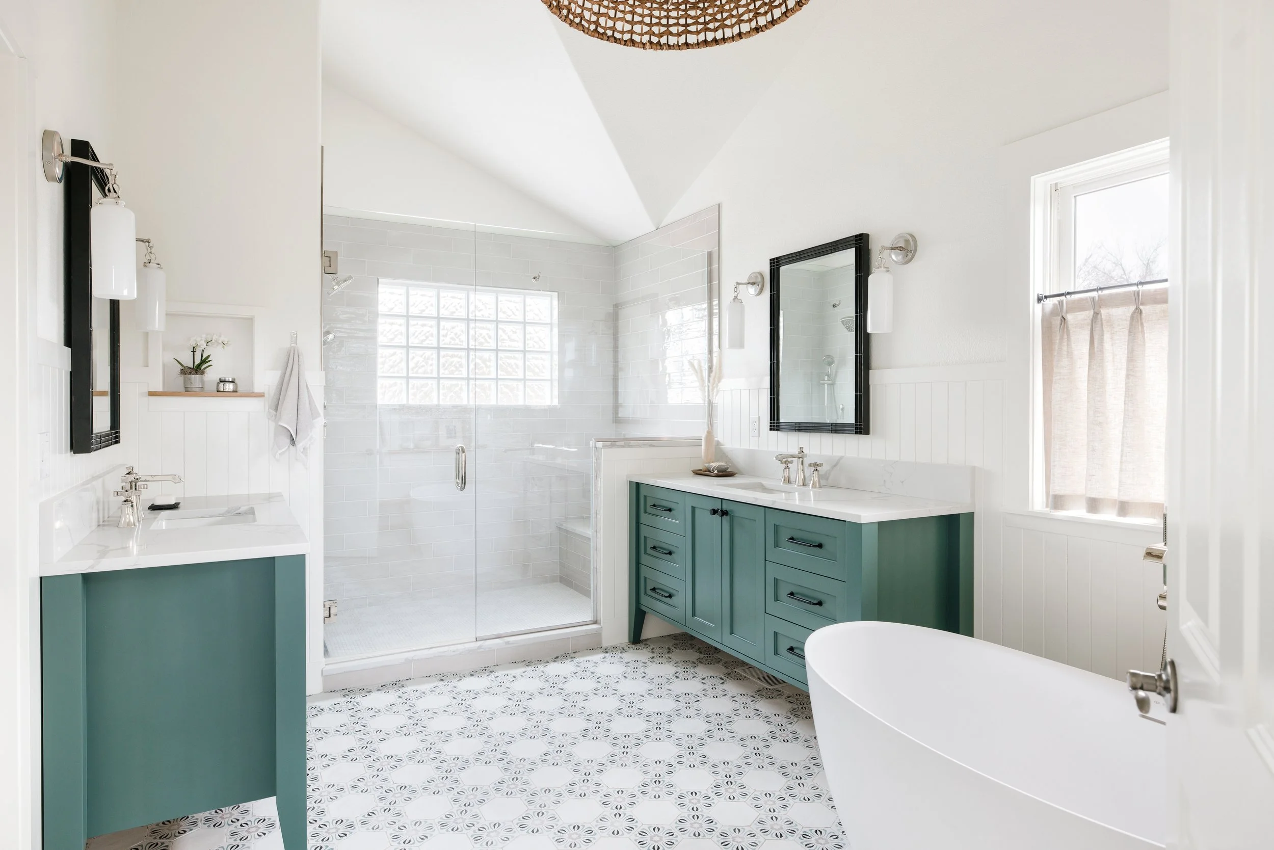 Modern bathroom with a white freestanding bathtub, teal vanity, glass-enclosed shower, black-framed mirror, and patterned tile floor.