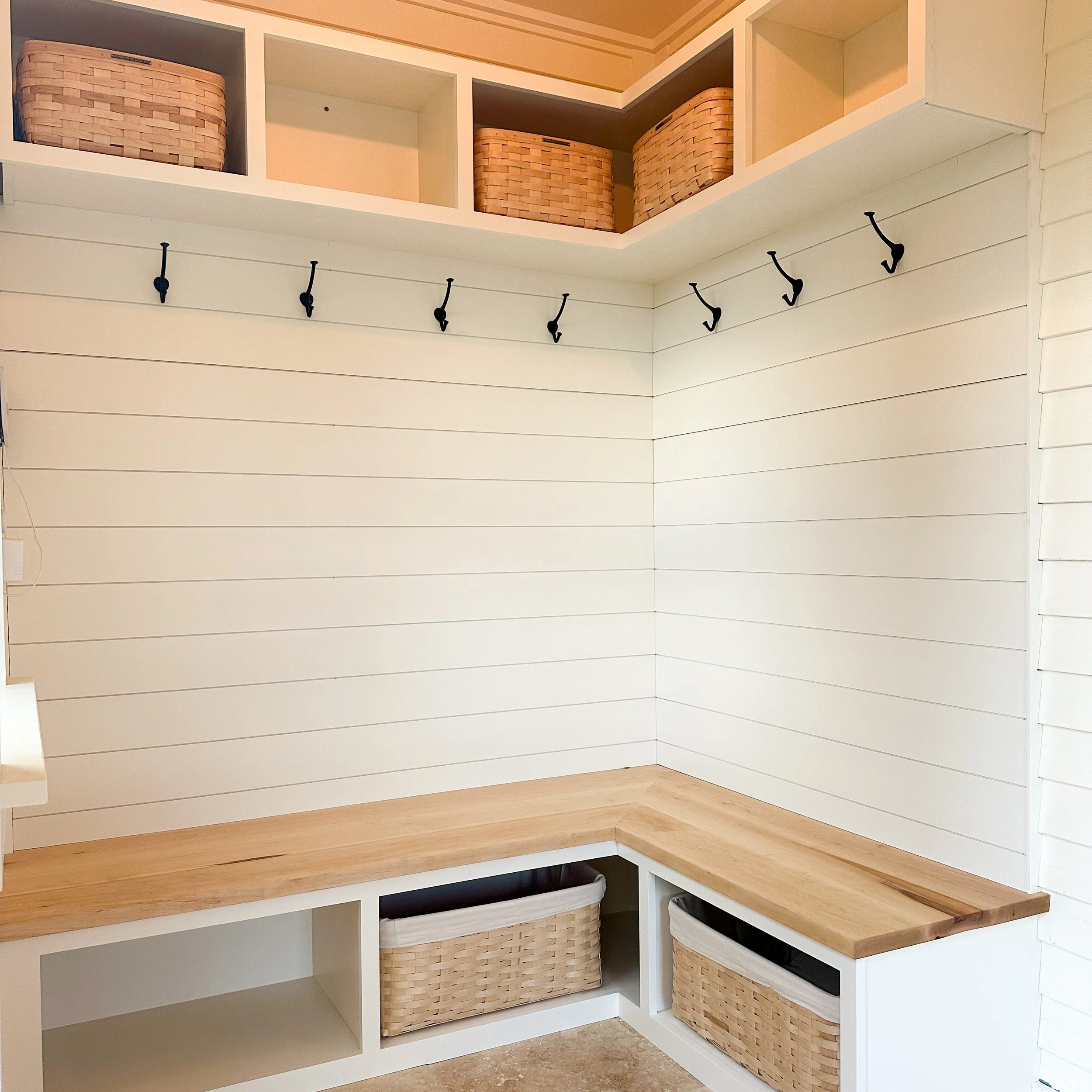 Corner of a room with white shiplap walls, a wooden bench with baskets underneath, and black hooks on the wall for hanging items.
