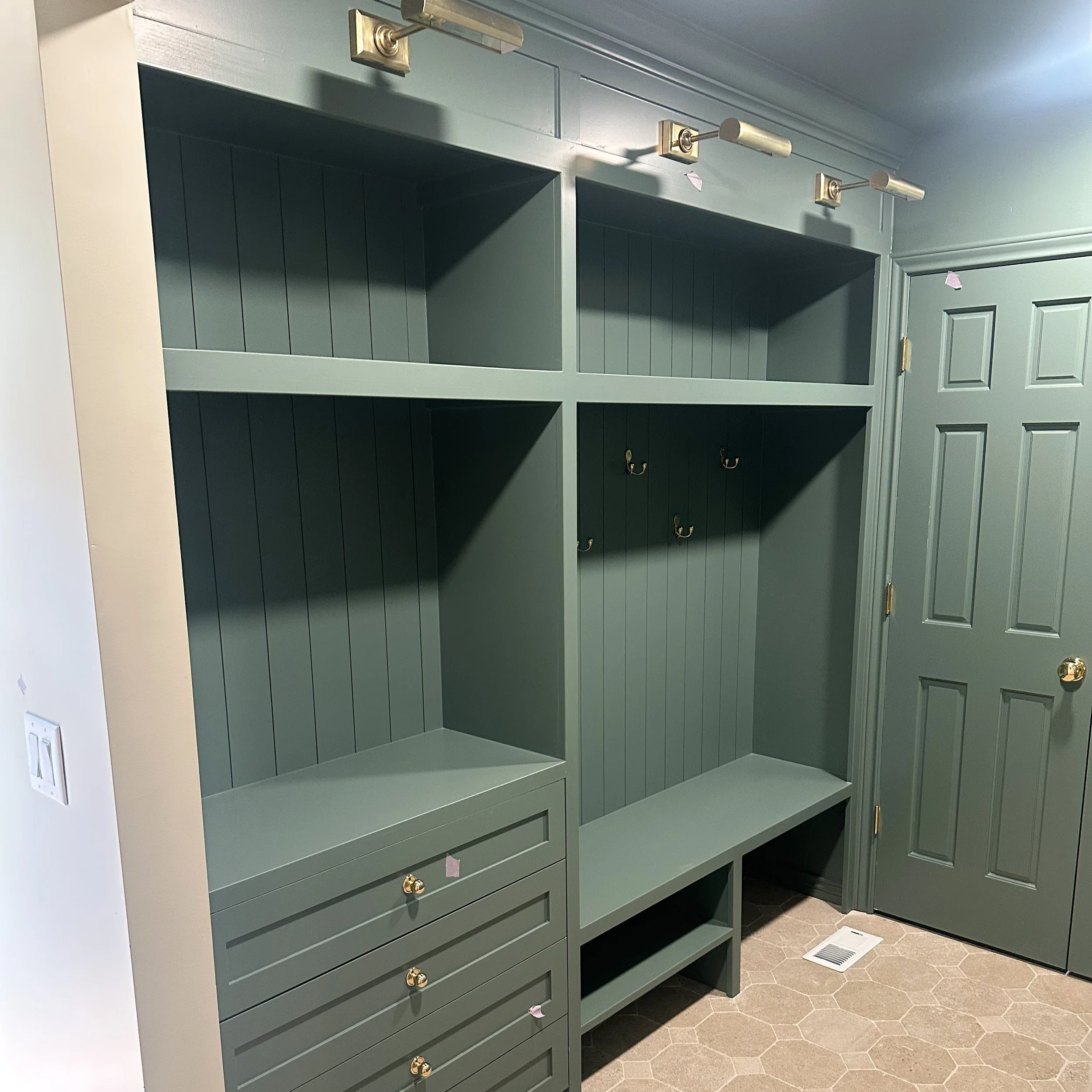Green built-in storage unit with open shelves, hooks, drawers with gold knobs, and a bench, adjacent to a green paneled door in a room with hexagonal tile flooring and a floor vent.