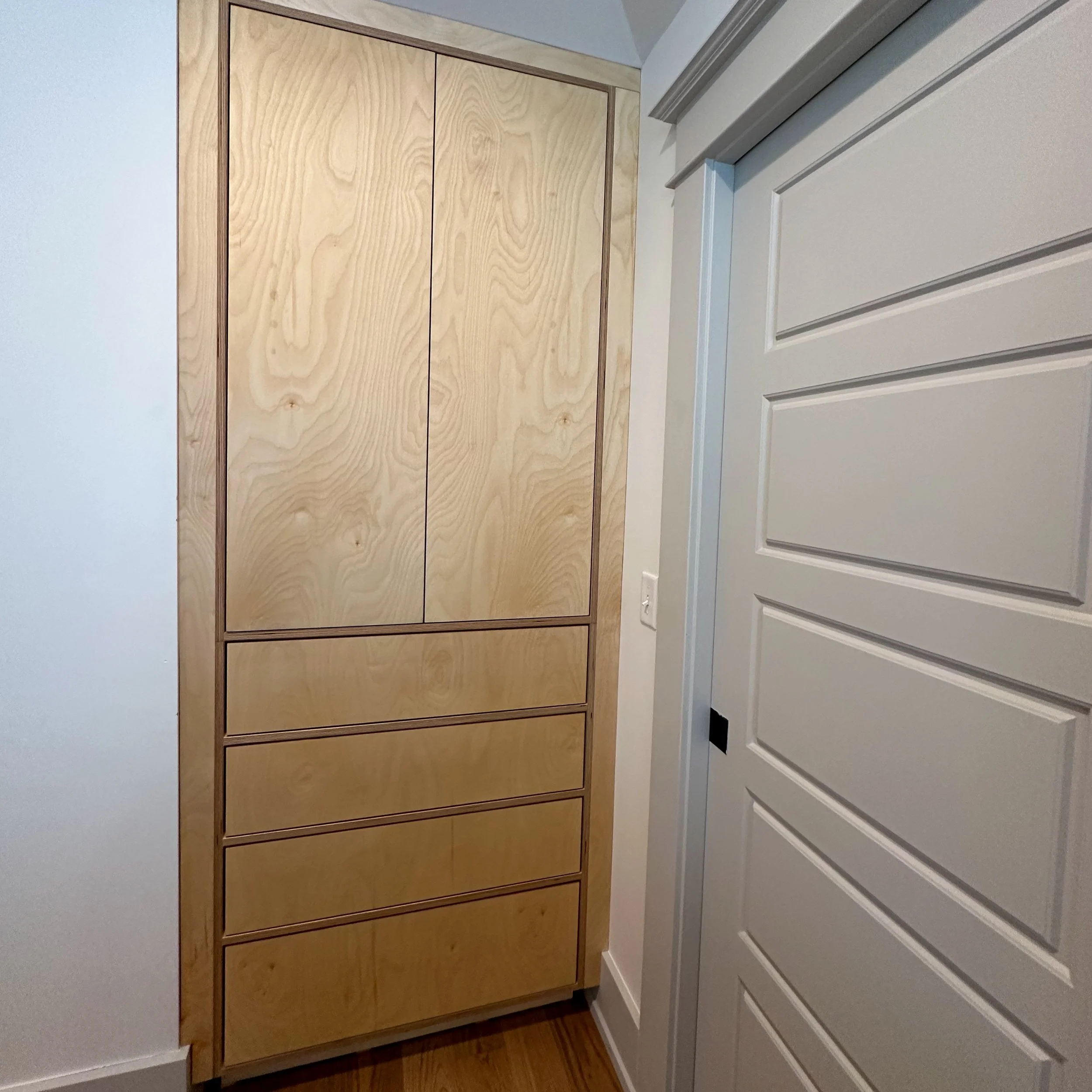 A tall wooden cabinet with six drawers and two upper doors, located in a corner next to a white paneled door in a room with white walls and a wooden floor.