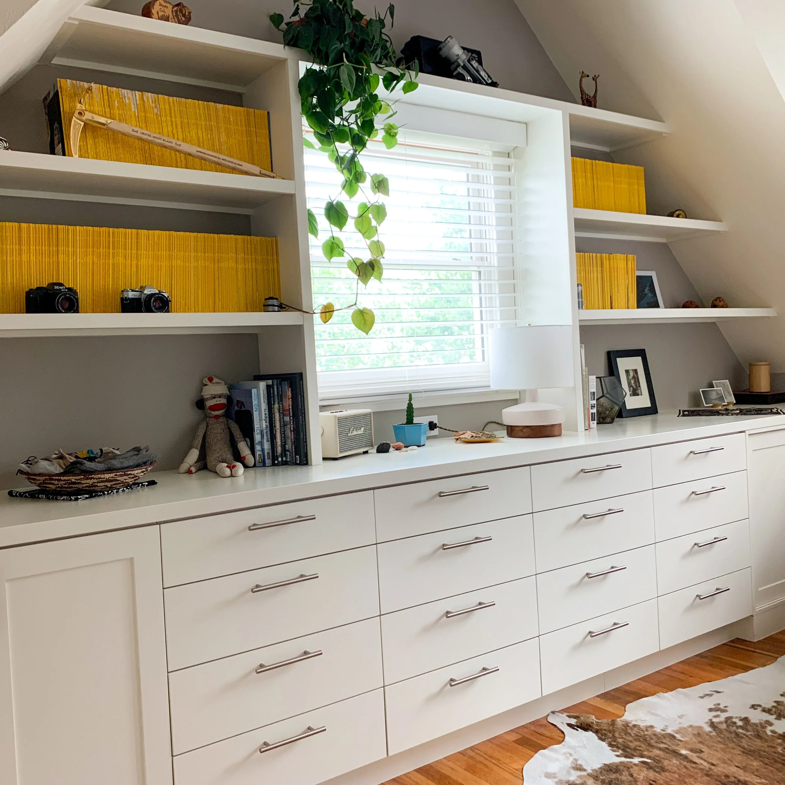 A white dresser with multiple drawers, decorated with various photo frames, a white table lamp, a small potted plant, and collectibles, with open shelves above holding books and decorative items, positioned below a window with blinds and a hanging pl