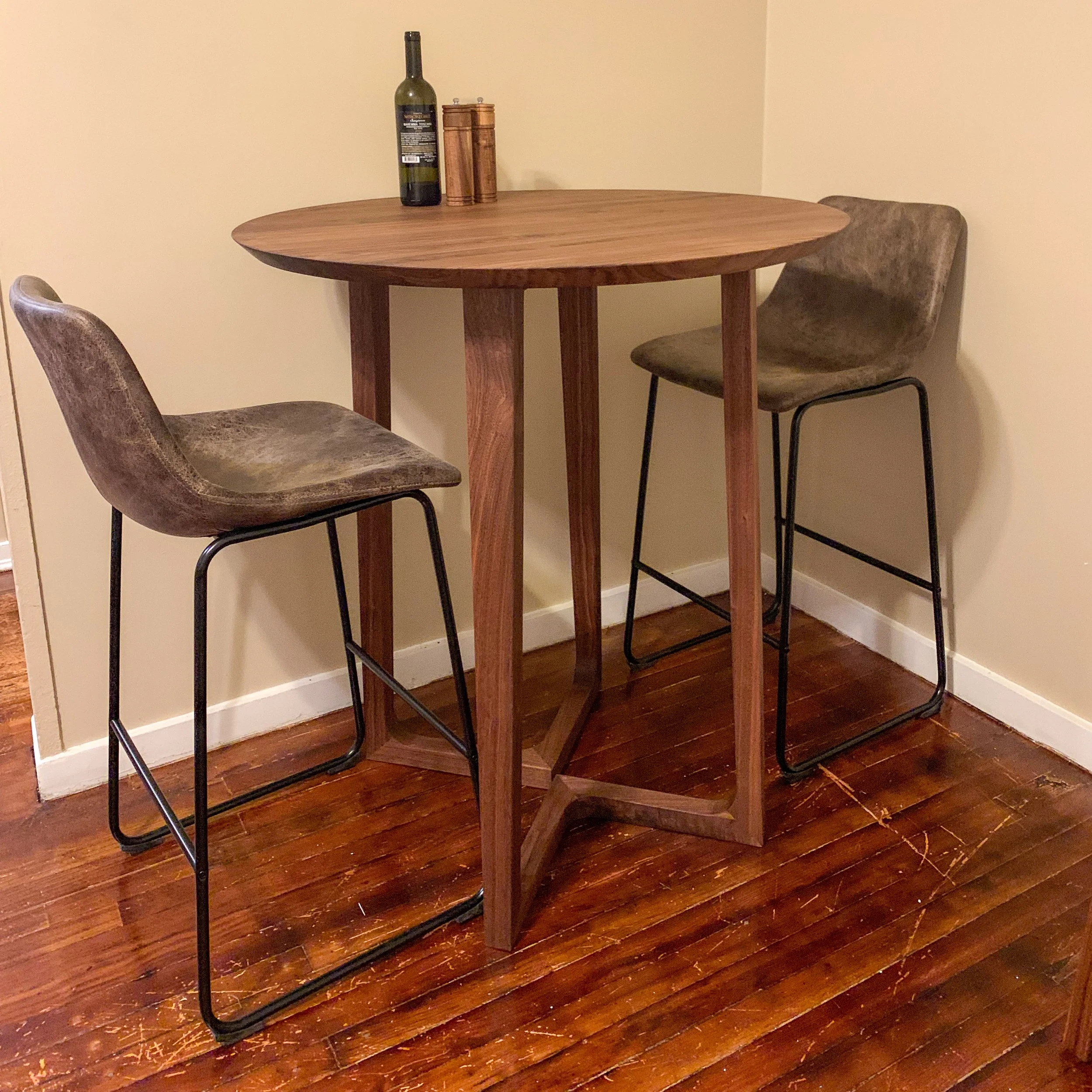 A round wooden table with two brown upholstered chairs with black metal legs, set against a plain light-colored wall, with a bottle of wine, two canisters, and a pepper mill on the table.