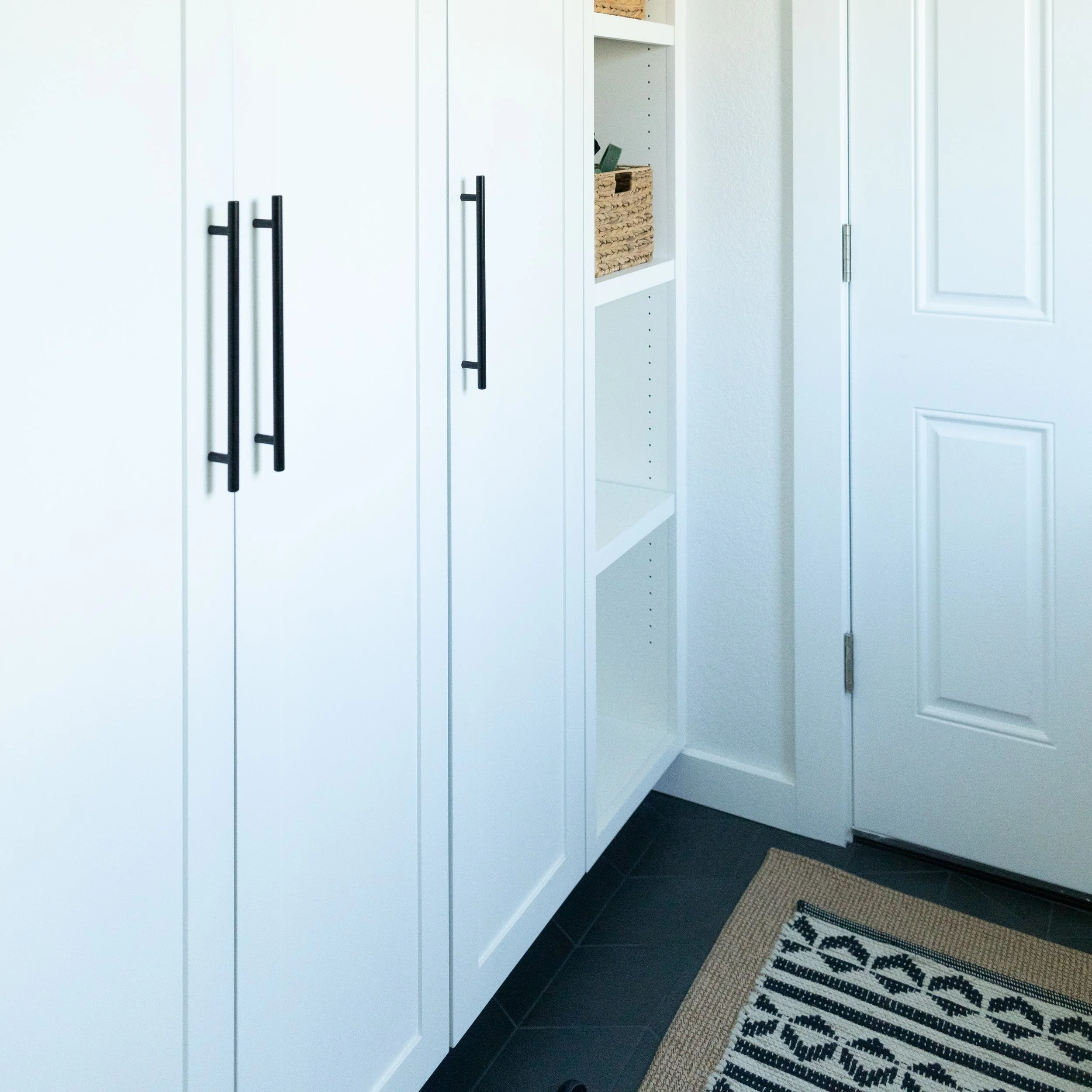 Close-up of white storage cabinets with black handles in an entryway next to a white door and a small open shelf with baskets and books, on a black tiled floor with a striped beige and black rug.