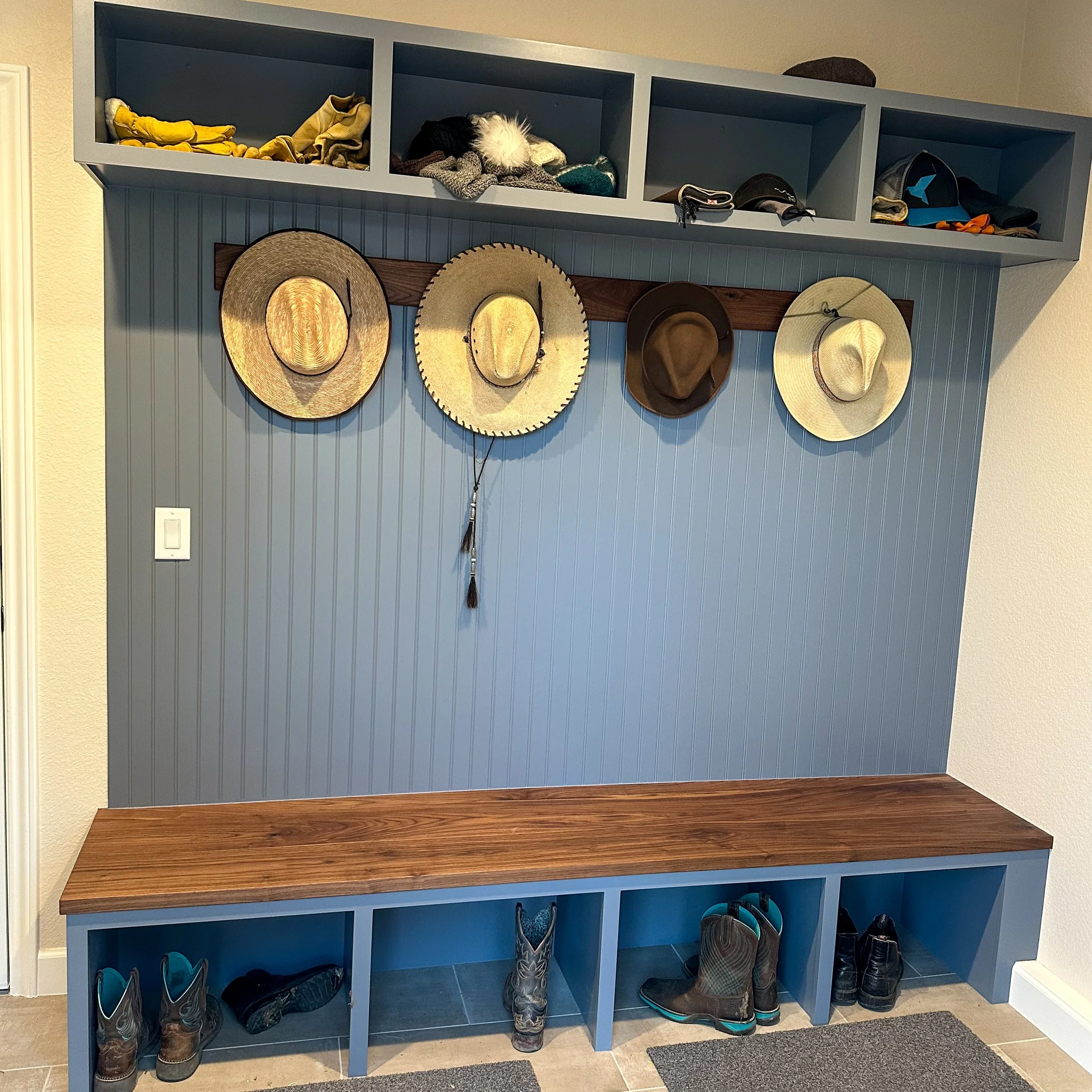 Entryway with a blue wall featuring four hanging hats, a row of shelves with shoes, boots, and hats, and a wood bench with cubbies for shoes below.