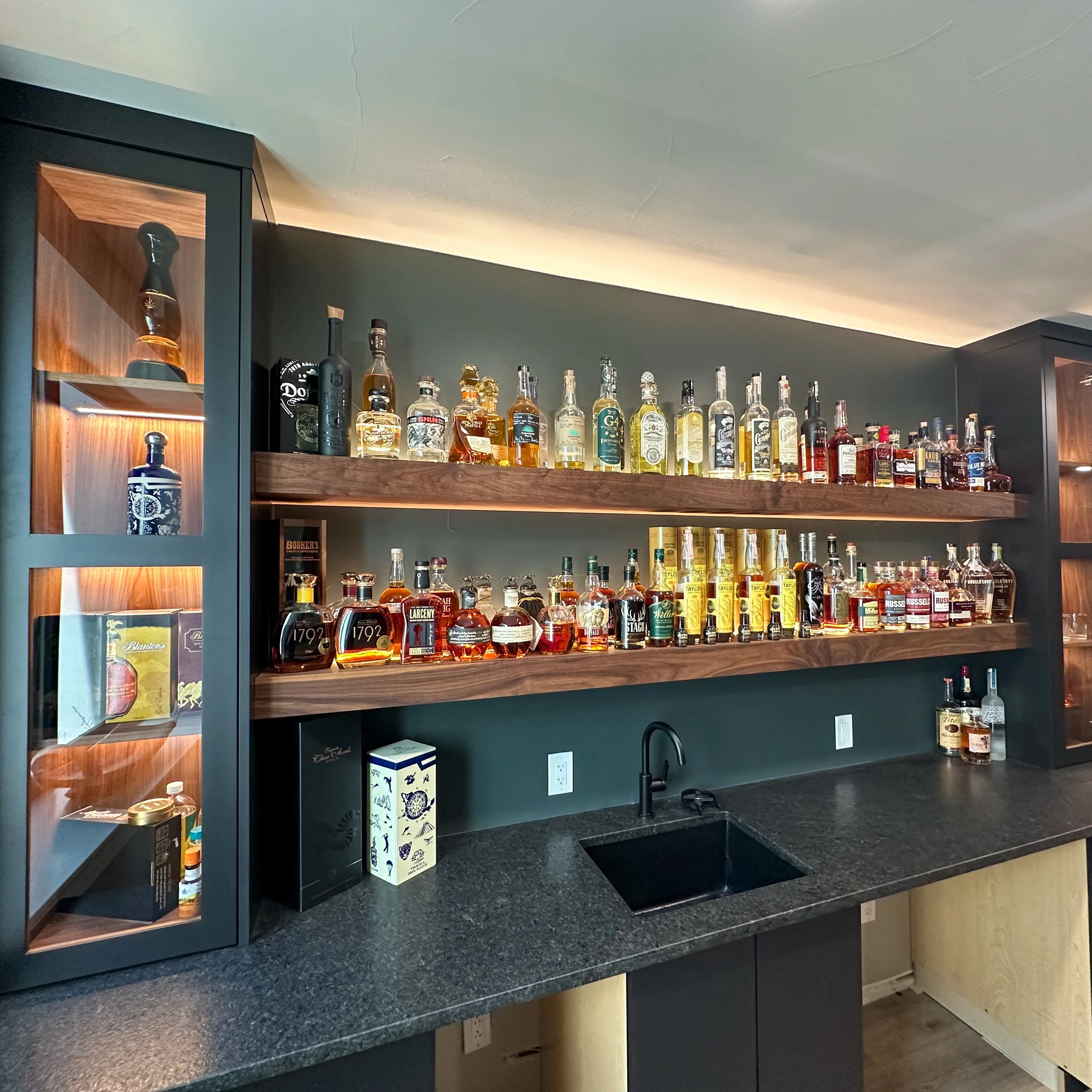 Bar with wooden shelves filled with various bottles of liquor, a black cabinet with a display of bottles inside, a black sink, and a dark countertop.