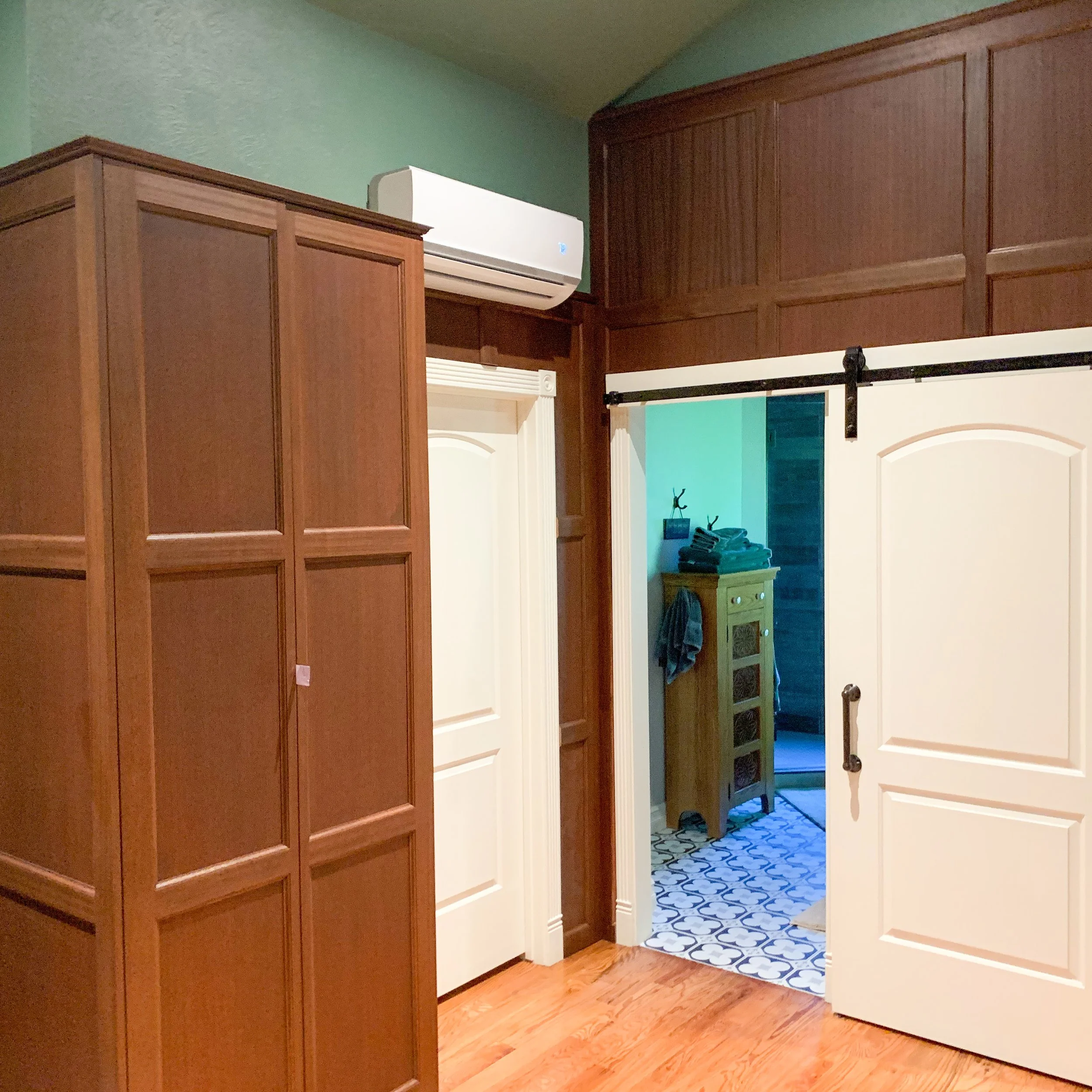 A room with wooden cabinets, white doors, a wall-mounted air conditioner, and an open sliding barn door revealing a hallway with a small green cabinet and patterned floor tiles.